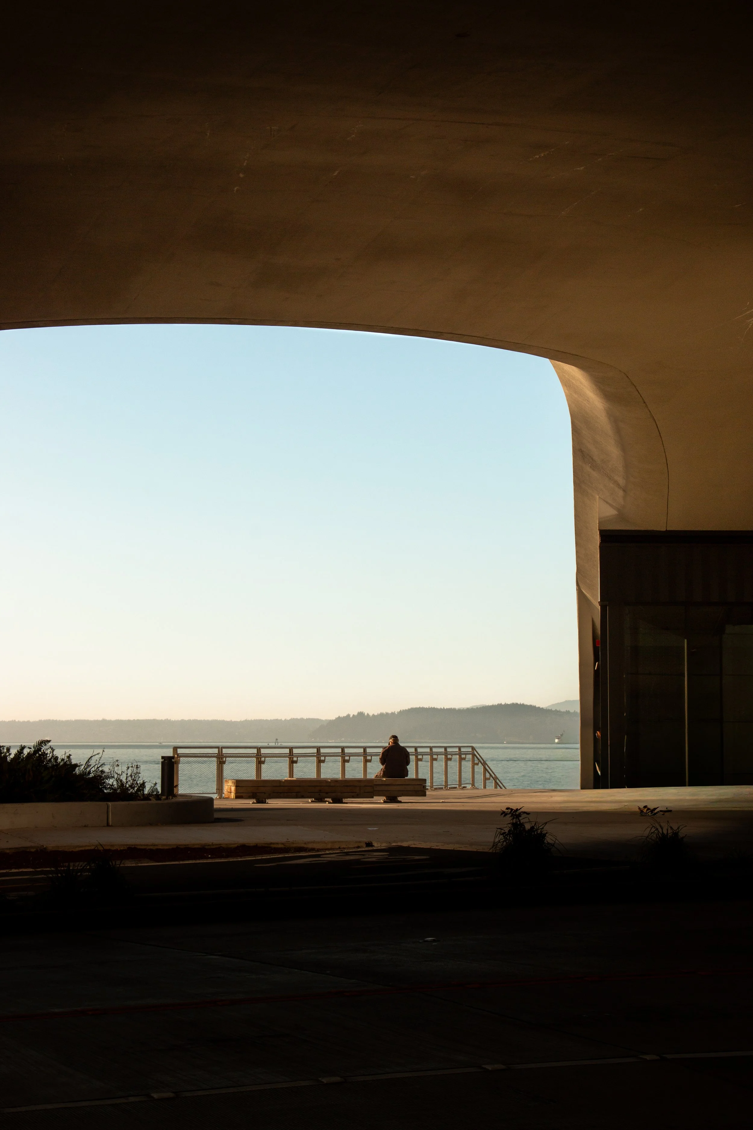 A person sitting on a bench looking out over a body of water, framed by a large architectural opening, during sunset or sunrise.