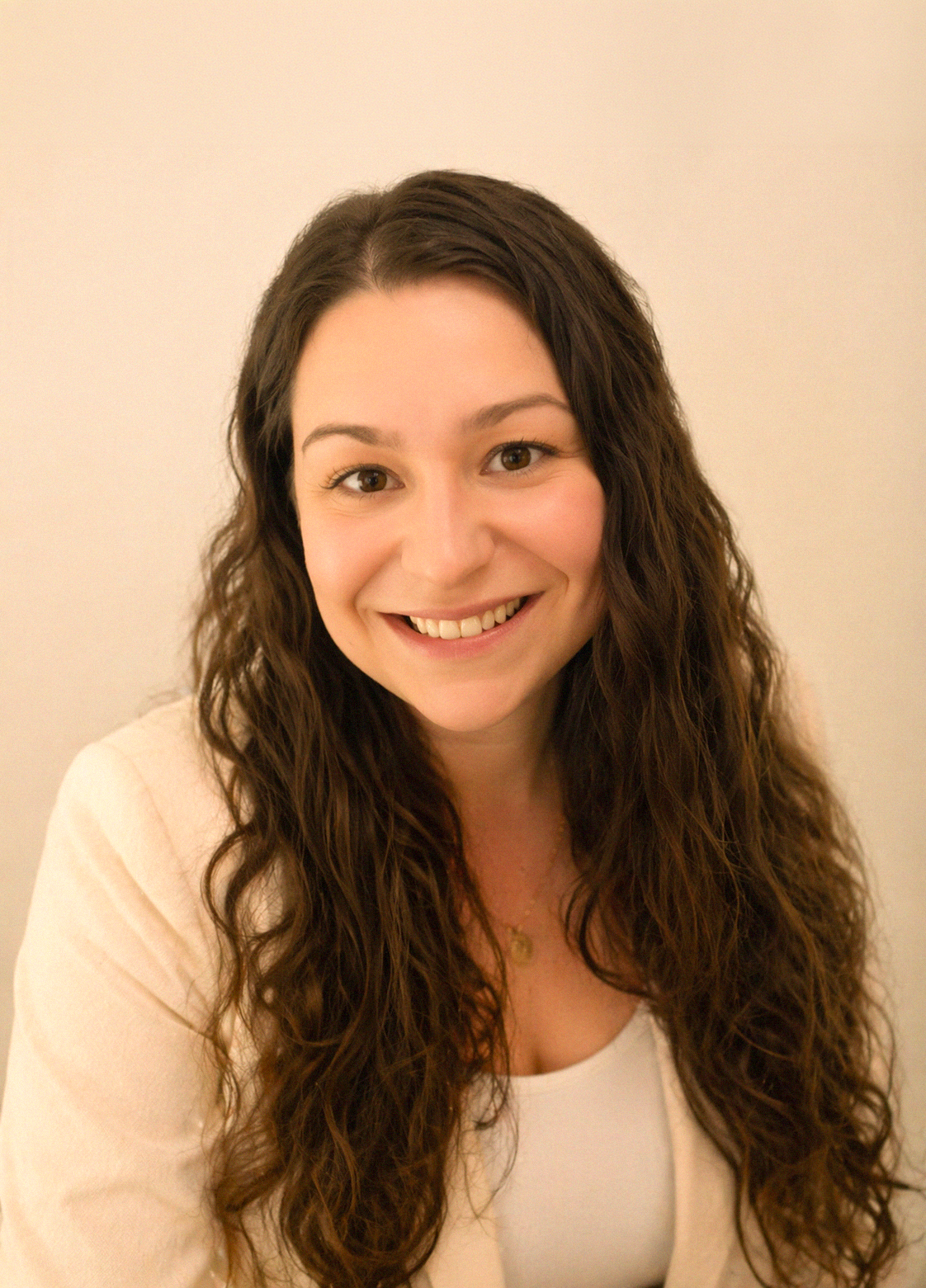 A woman with long, curly brown hair smiling at the camera against a plain, light-colored background.