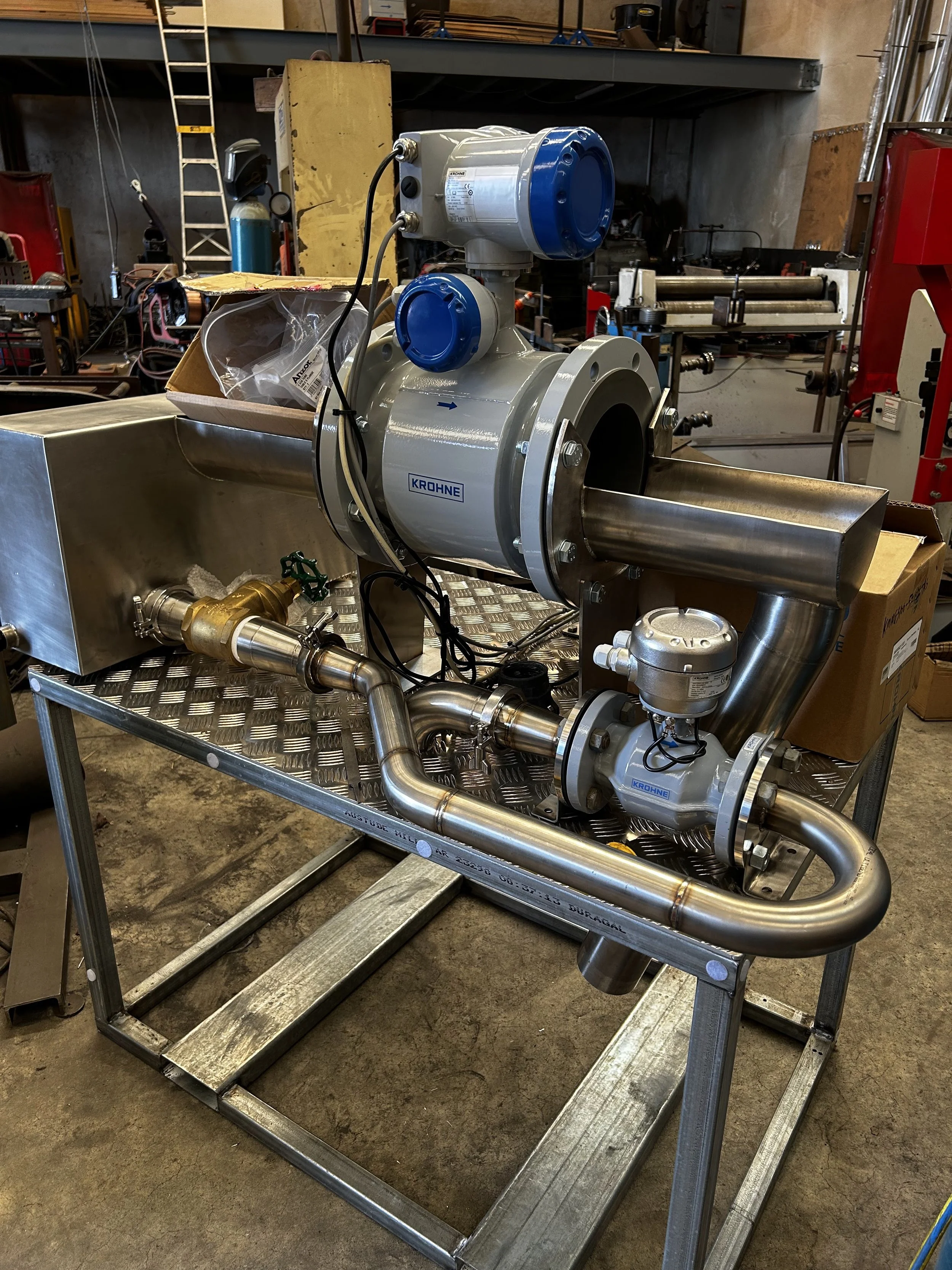 A metal industrial pump with pipes, mounted on a metal frame, in a workshop setting. Various tools and equipment visible in the background.