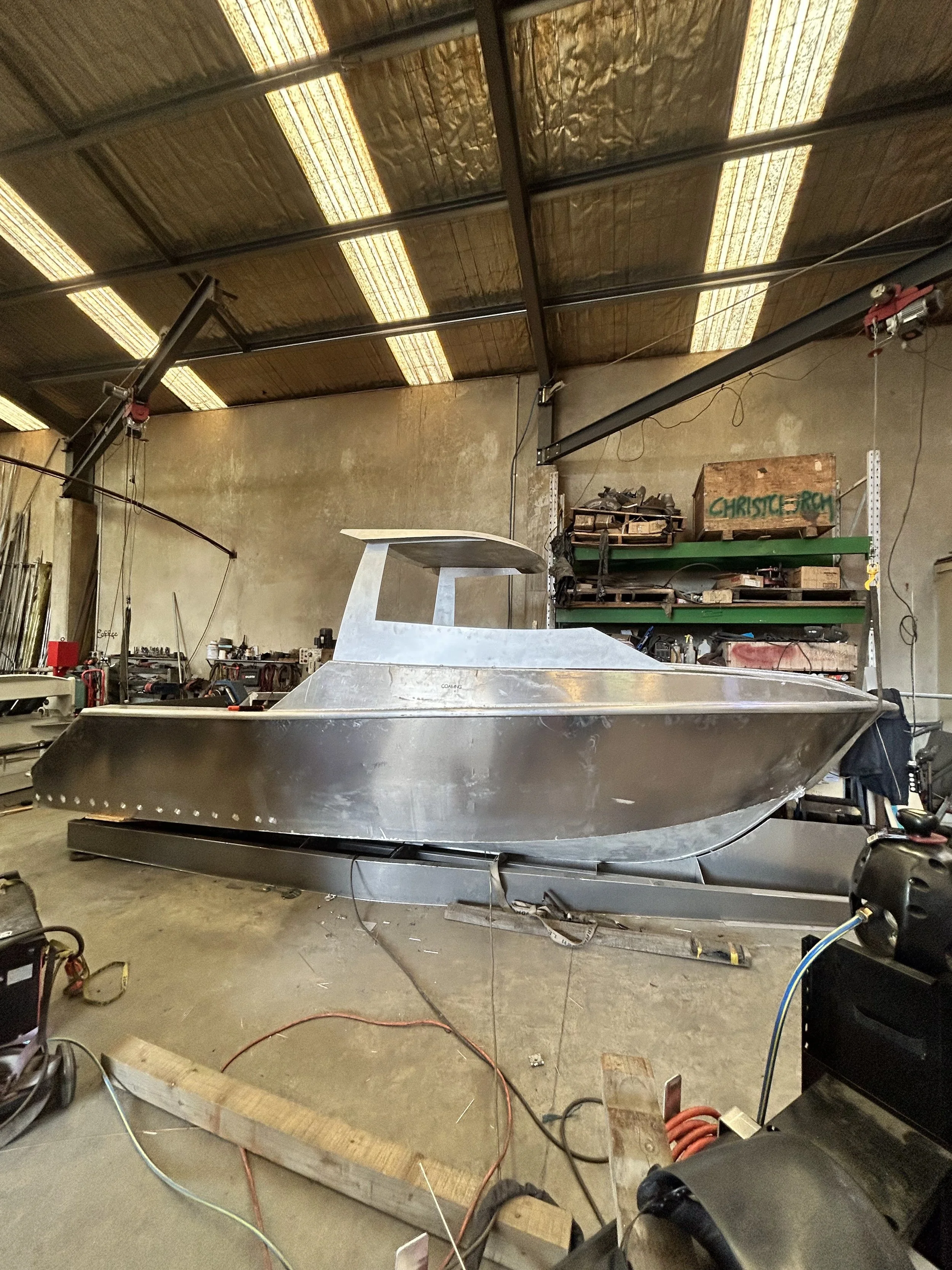 A workshop with a metal boat under construction, various tools and equipment around, and shelves with boxes and other items, in a spacious industrial space with a high ceiling.