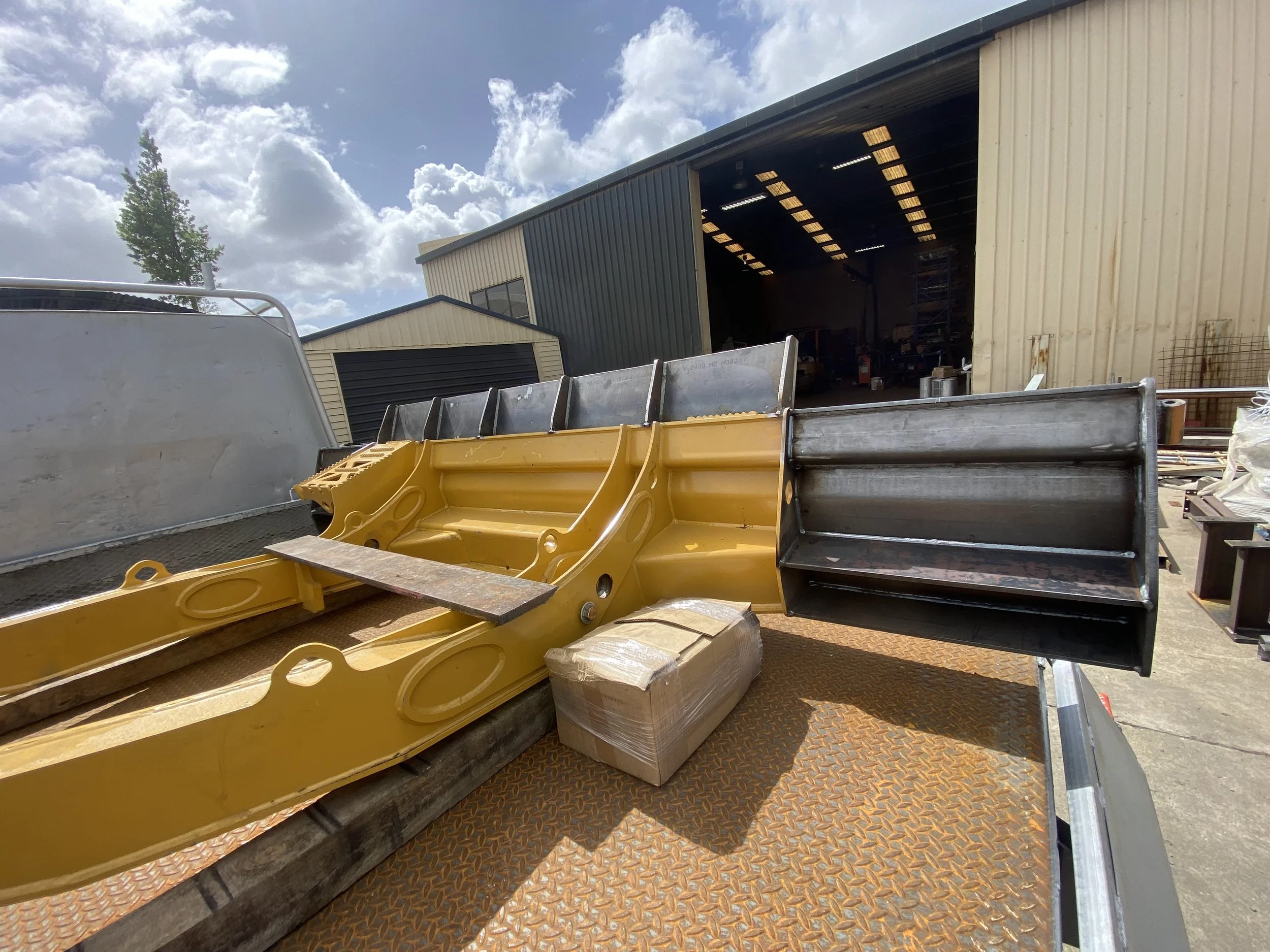 View of an open garage or workshop with various construction tools and equipment, including yellow safety barriers and a large container, with a metal building and a person inside in the background under a cloudy sky.