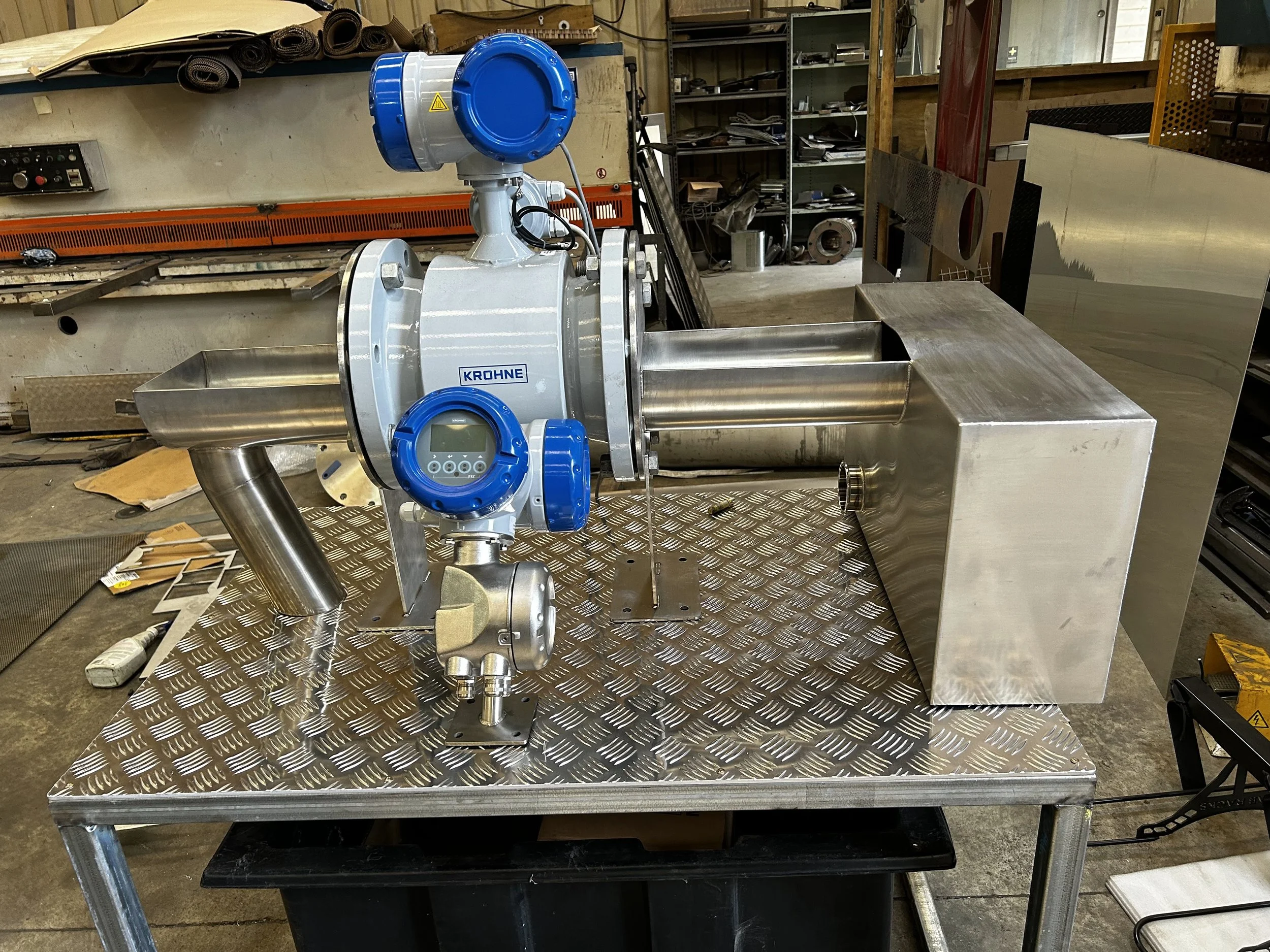 A stainless steel machine with a digital control panel labeled 'KROHNE' installed on a metal work table in a workshop. The workshop has shelves with various tools and materials, and there are pieces of metal and other equipment scattered around.