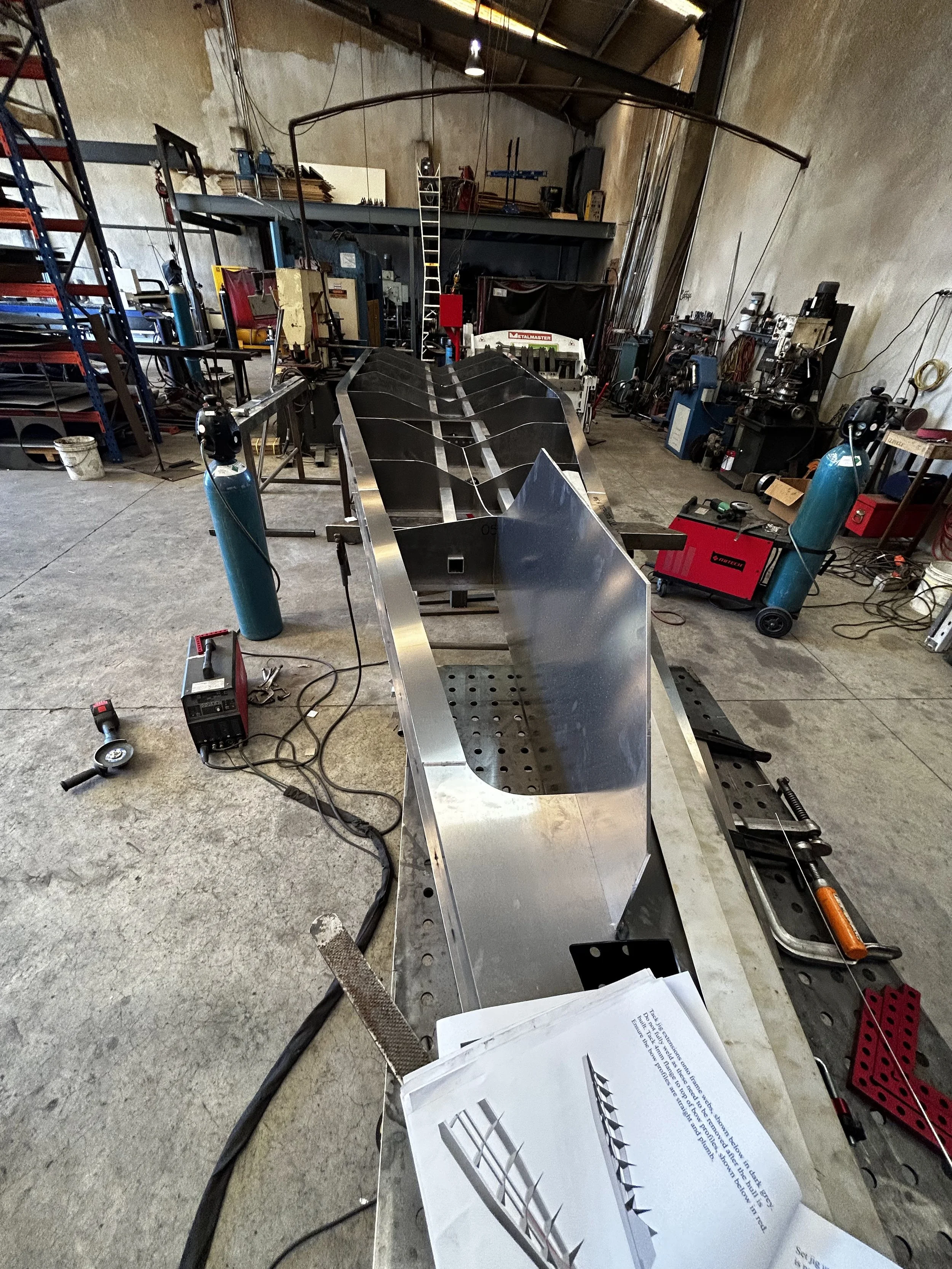 A workshop with metalworking equipment, including an aluminum boat hull in progress, various tools, and a workbench with diagrams and technical plans.
