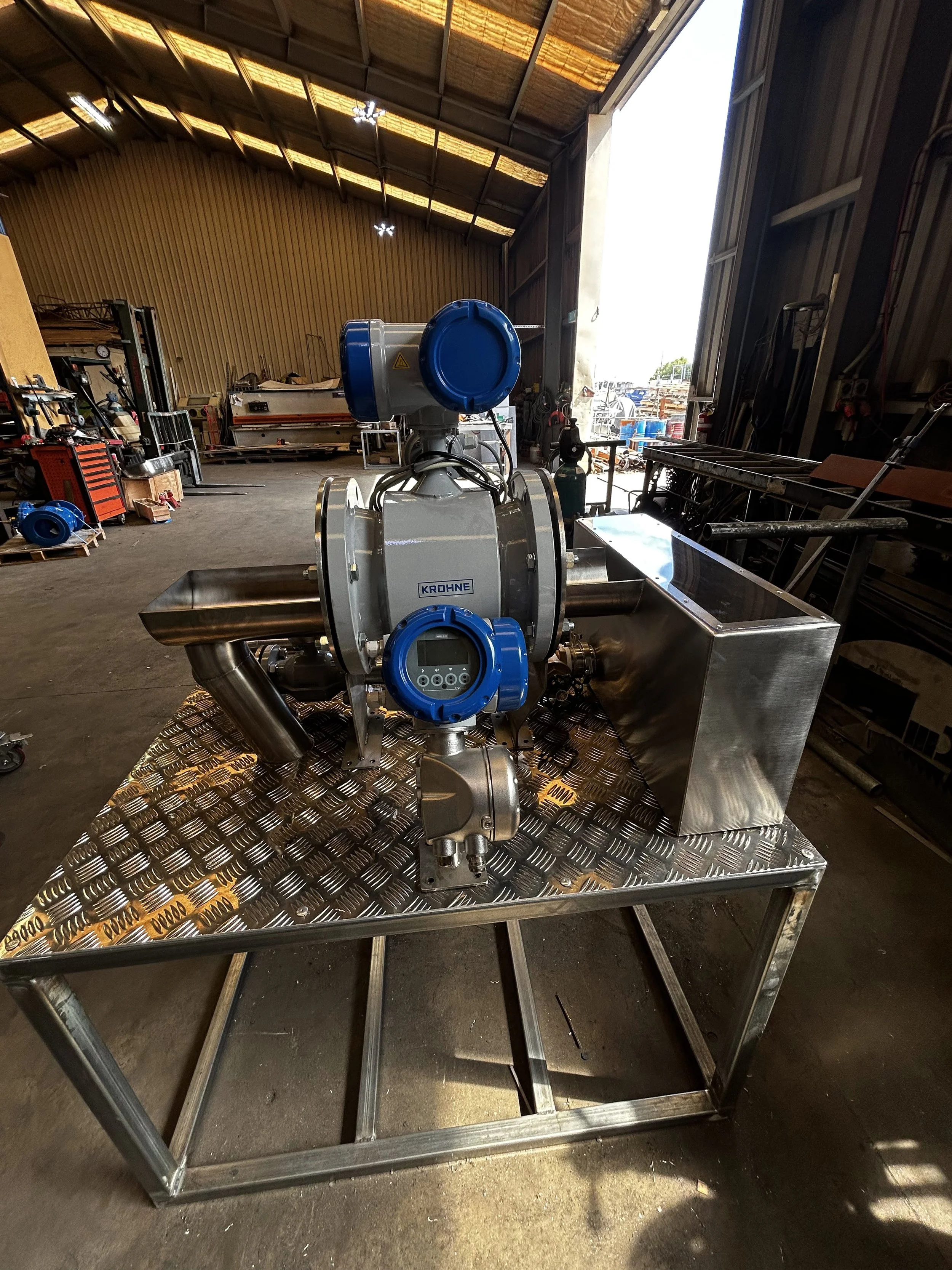 Industrial workshop with machinery and equipment, including stainless steel tanks and tools, inside a large warehouse with a high ceiling.