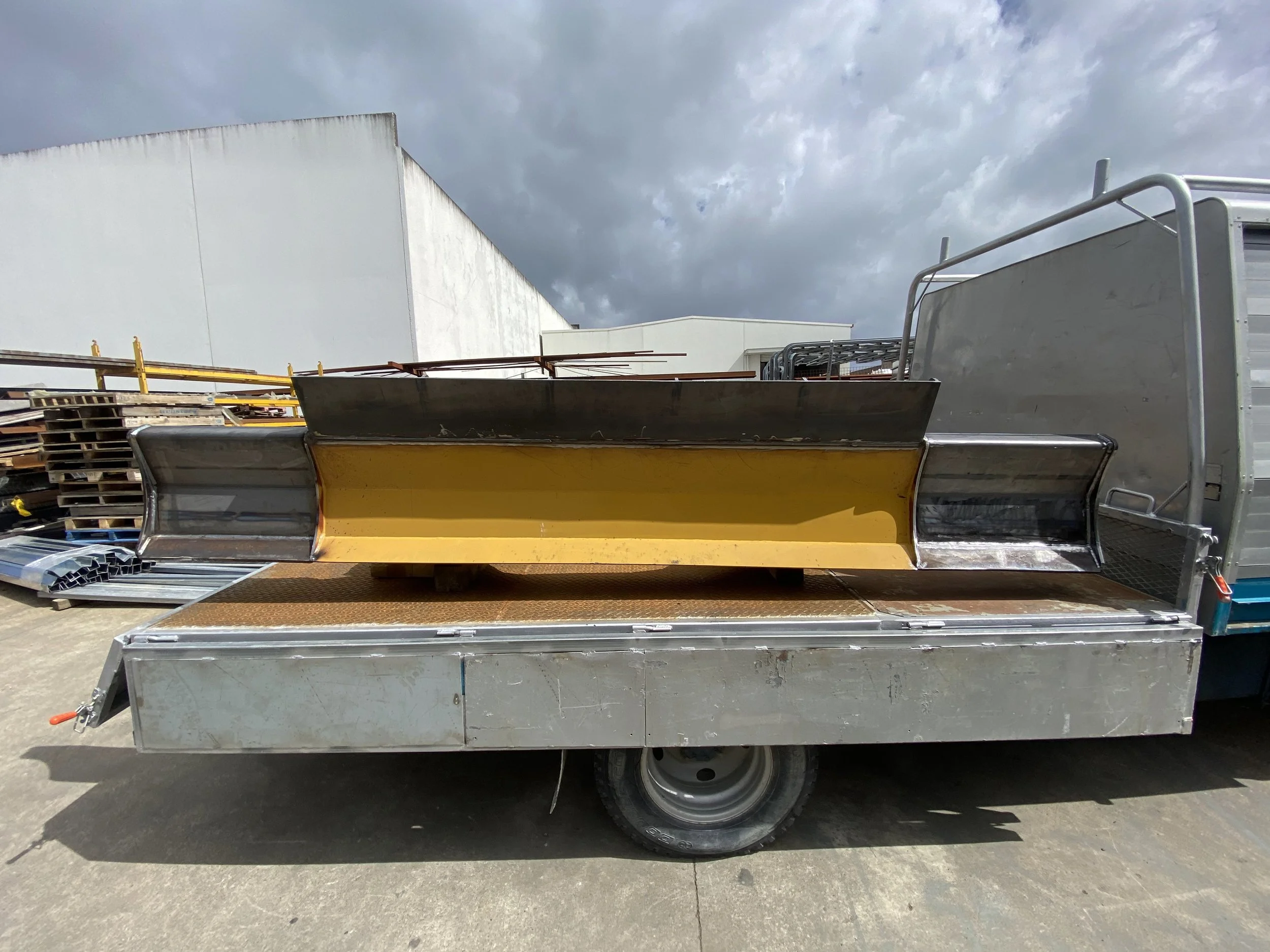 A flatbed truck carrying various metal and wooden construction materials, including a yellow snow plow blade, against a cloudy sky.