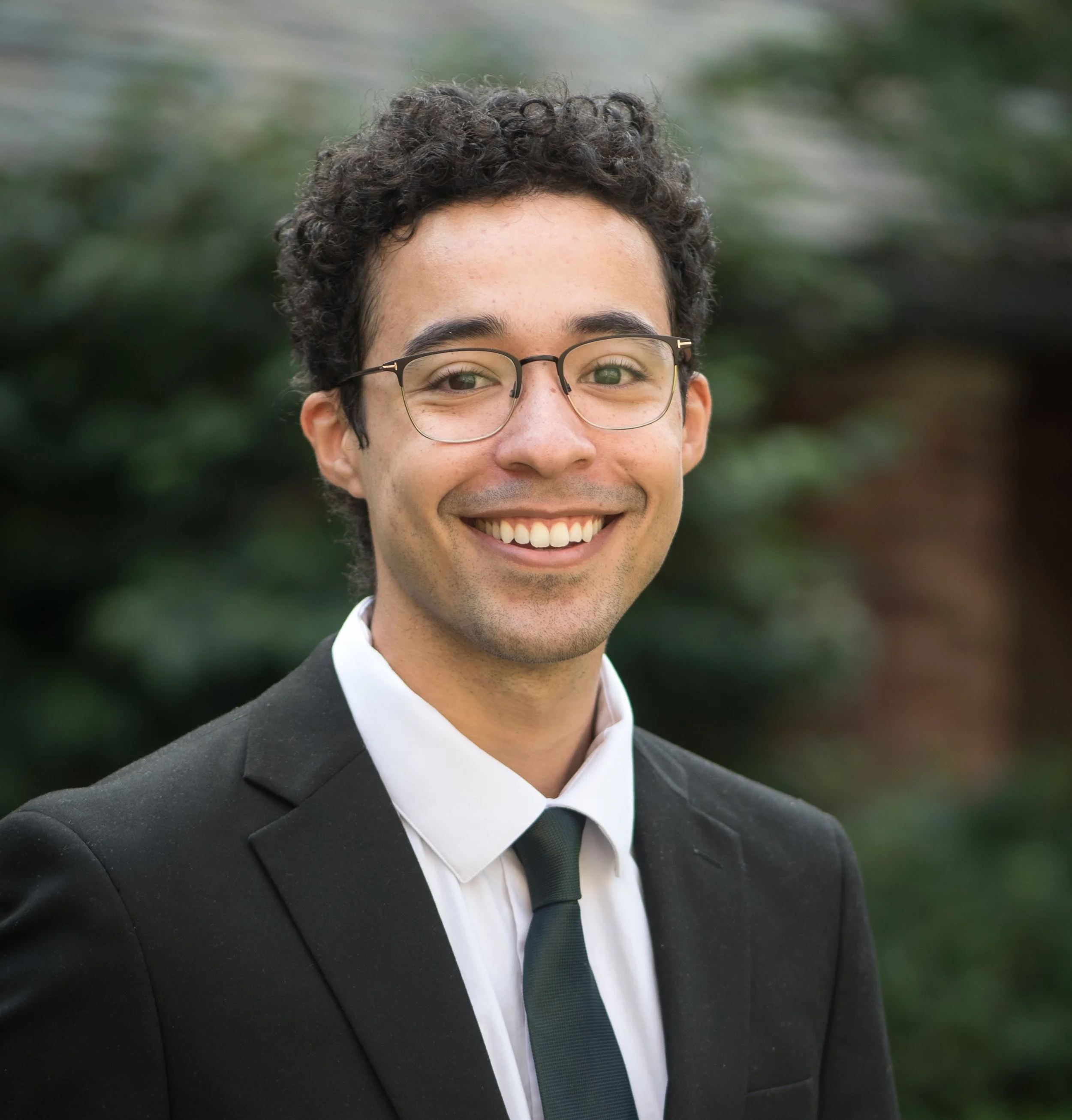 Zachary Williams Yale Law School. Zachary Williams Yale Law. Zachary Williams Yale. A smiling young man with curly dark hair, glasses, wearing a black suit, white shirt, and black tie, outdoors with blurred greenery in the background.