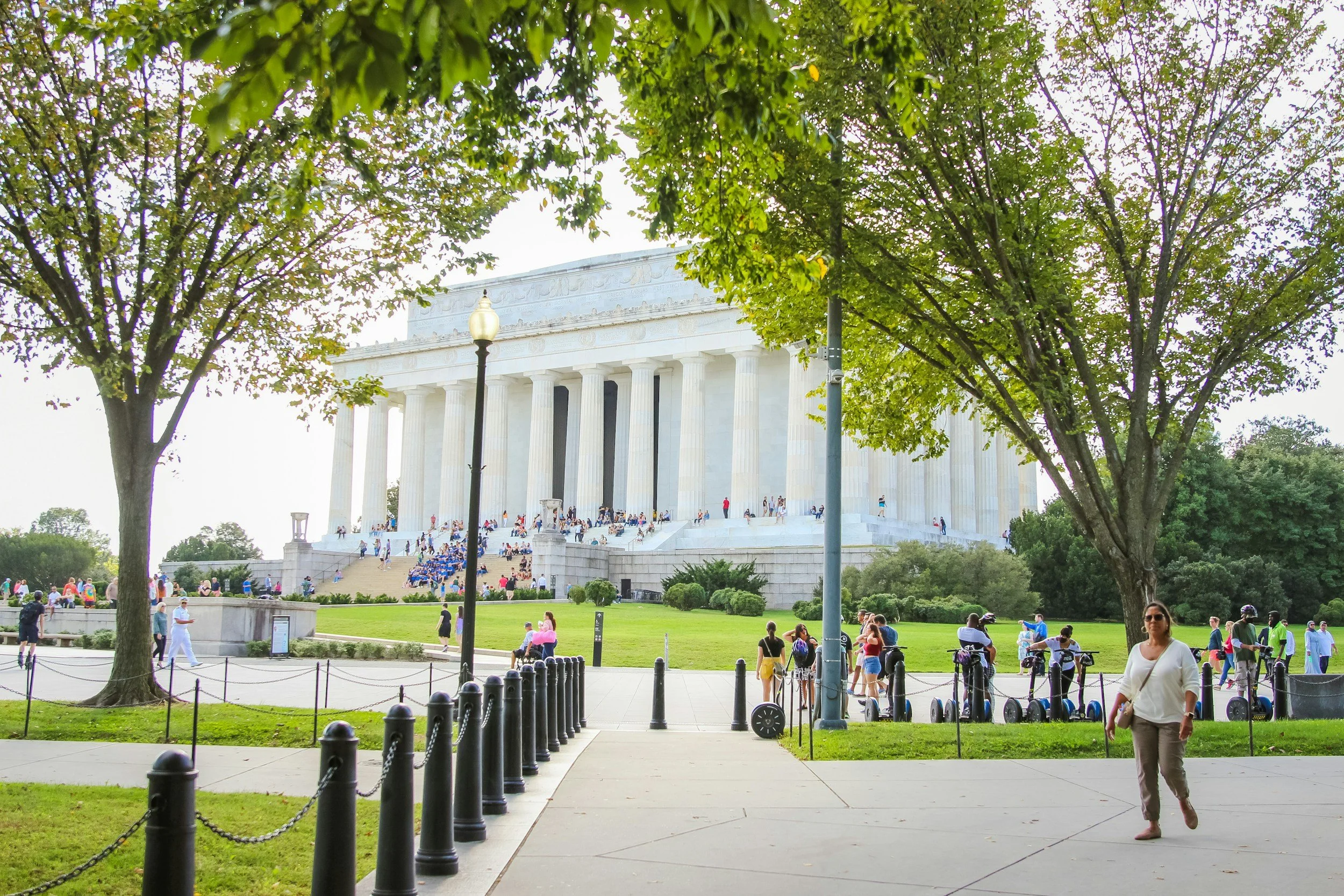 People visiting the Lincoln Memorial in Washington, D.C., with large trees in the foreground and a clear sky.