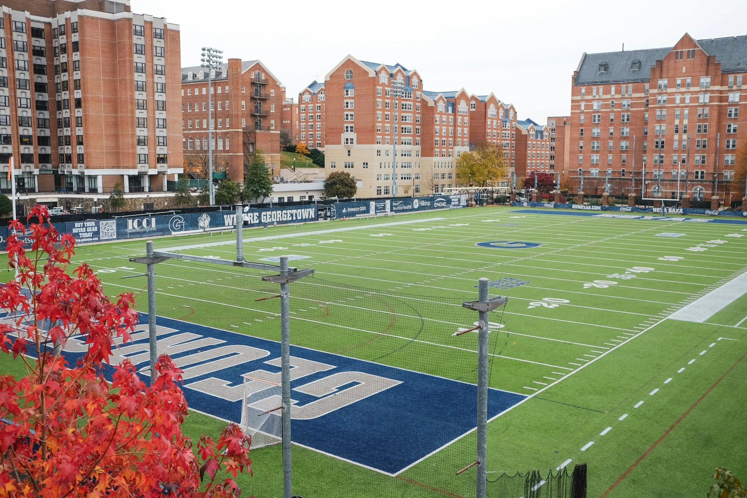 A football field with vibrant green turf, marked with white yard lines and blue end zones, is situated in front of several multi-story red brick residential buildings. Red autumn leaves are visible in the foreground.
