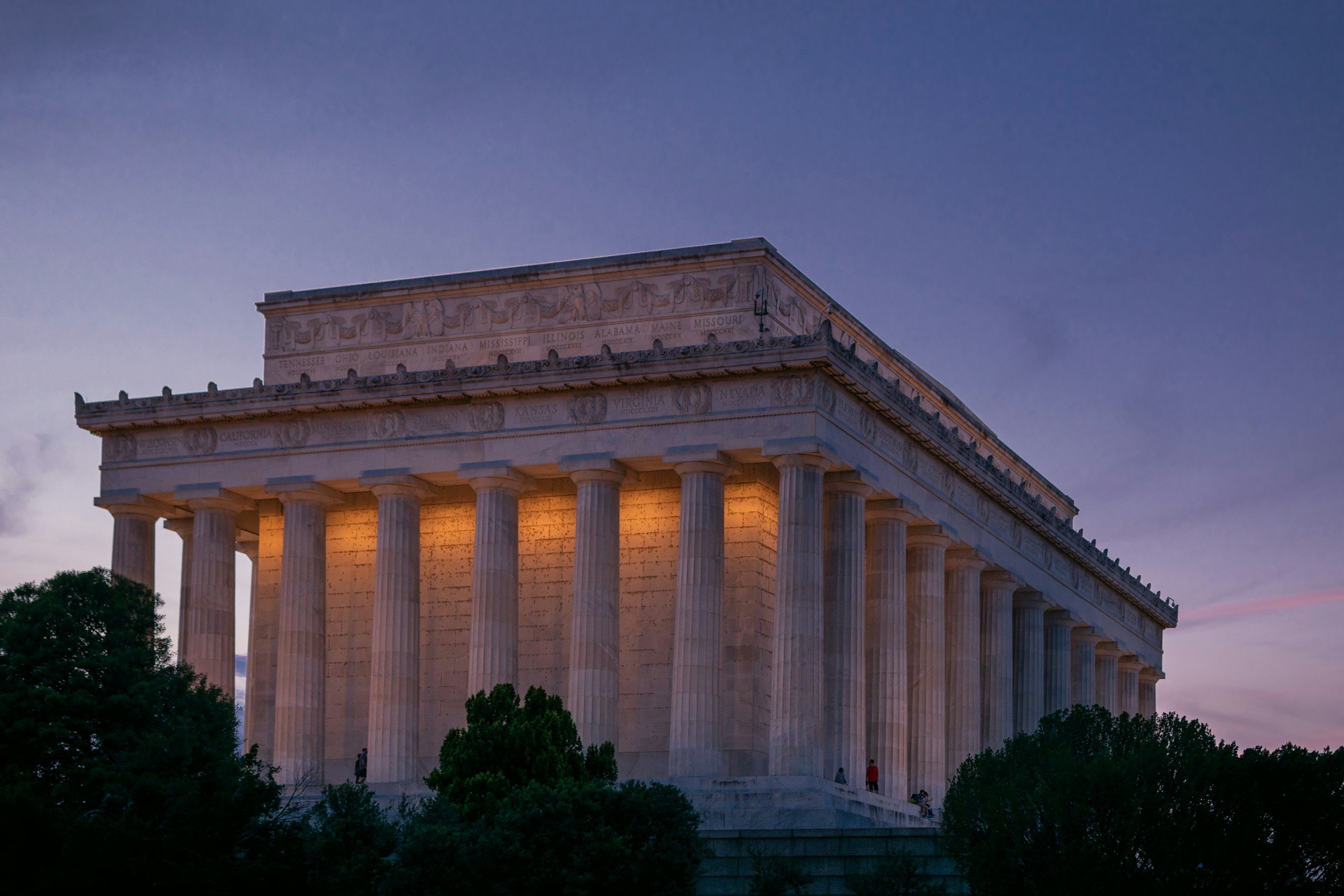 The Lincoln Memorial illuminated at dusk with a clear sky and trees in the foreground.