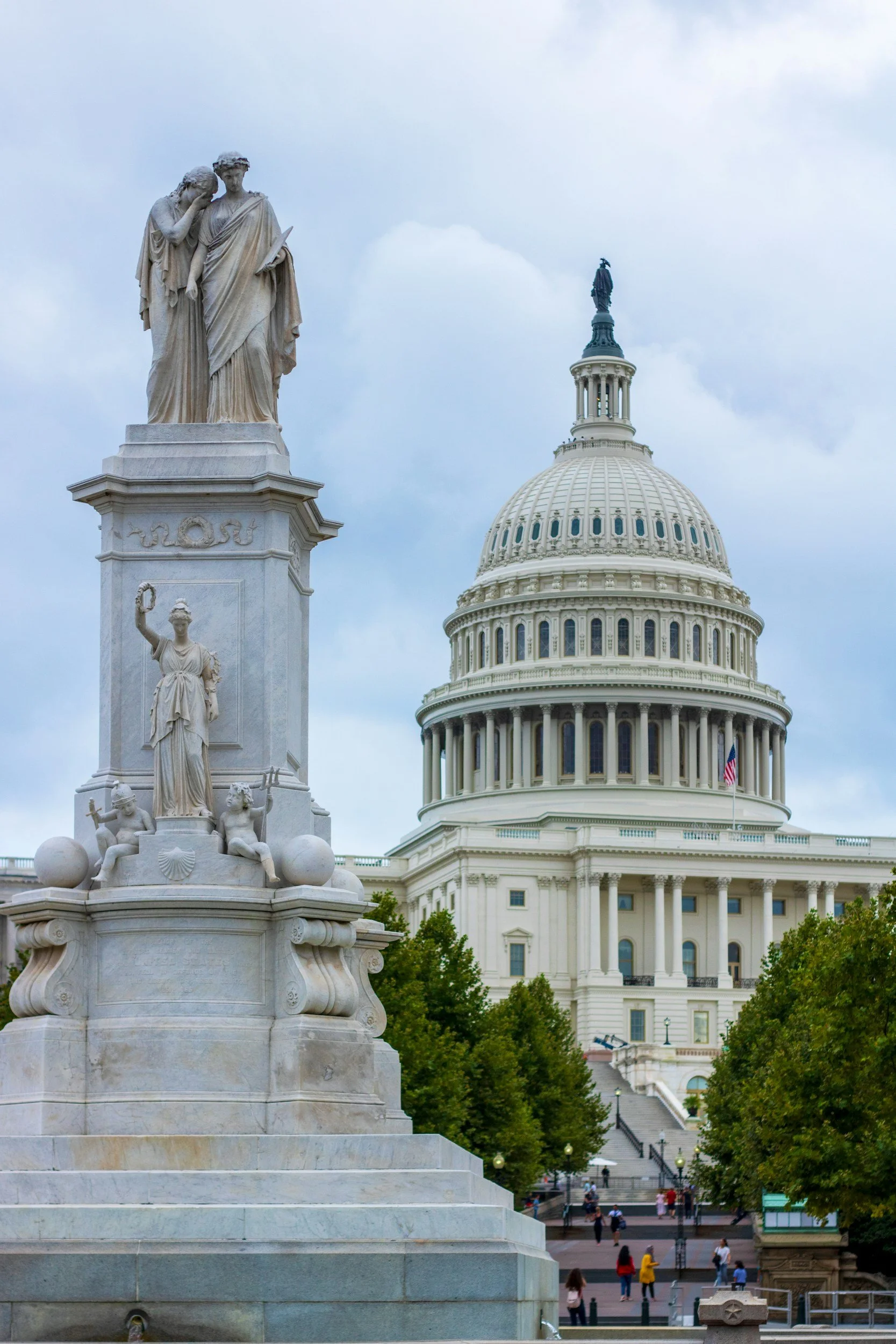 View of the United States Capitol building with its iconic dome and a nearby monument with statues in the foreground. Several people are walking and gathering in the park area.
