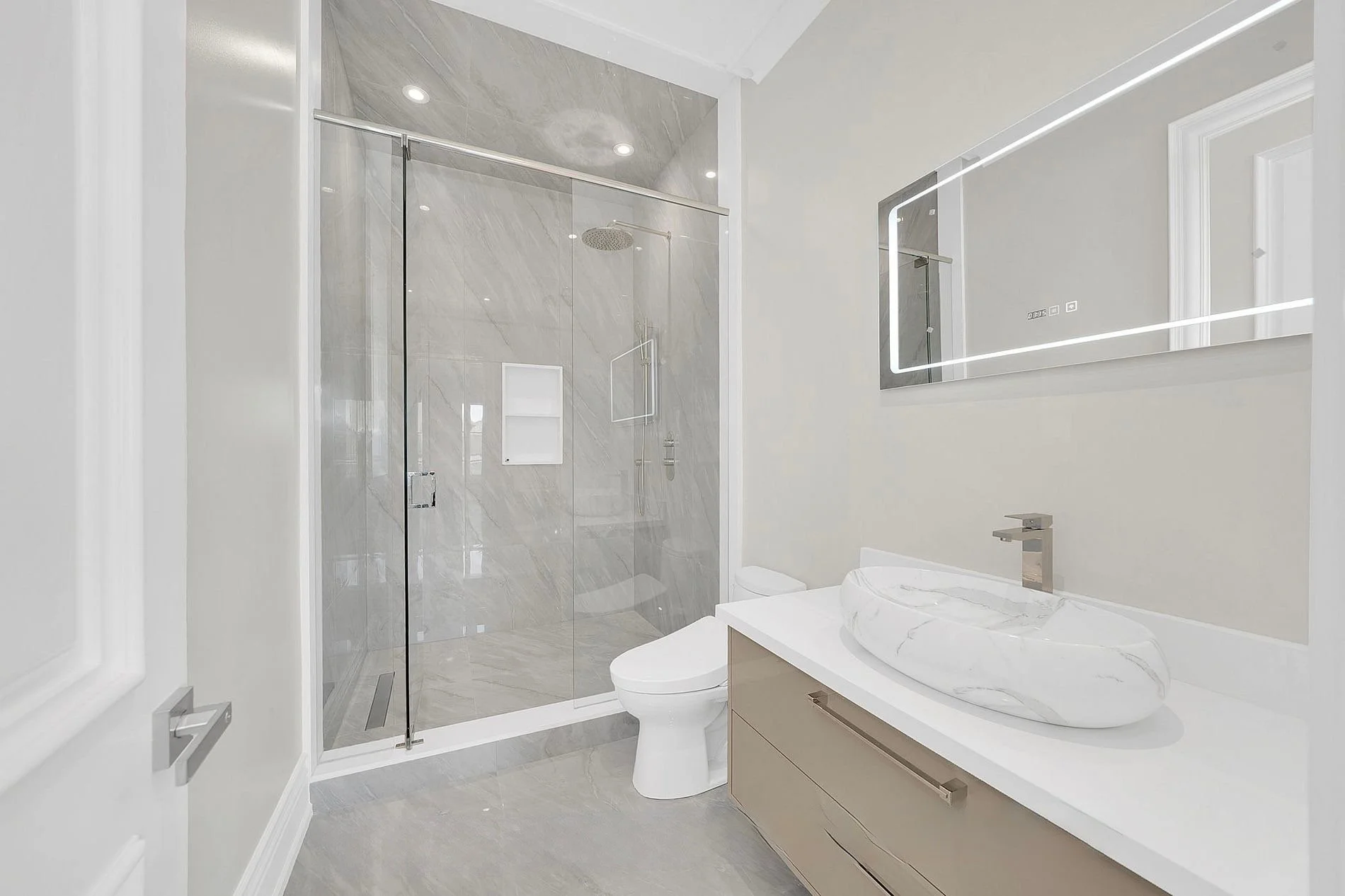 Modern bathroom with a walk-in shower, a white toilet, beige vanity with a marble vessel sink, and a large illuminated mirror.
