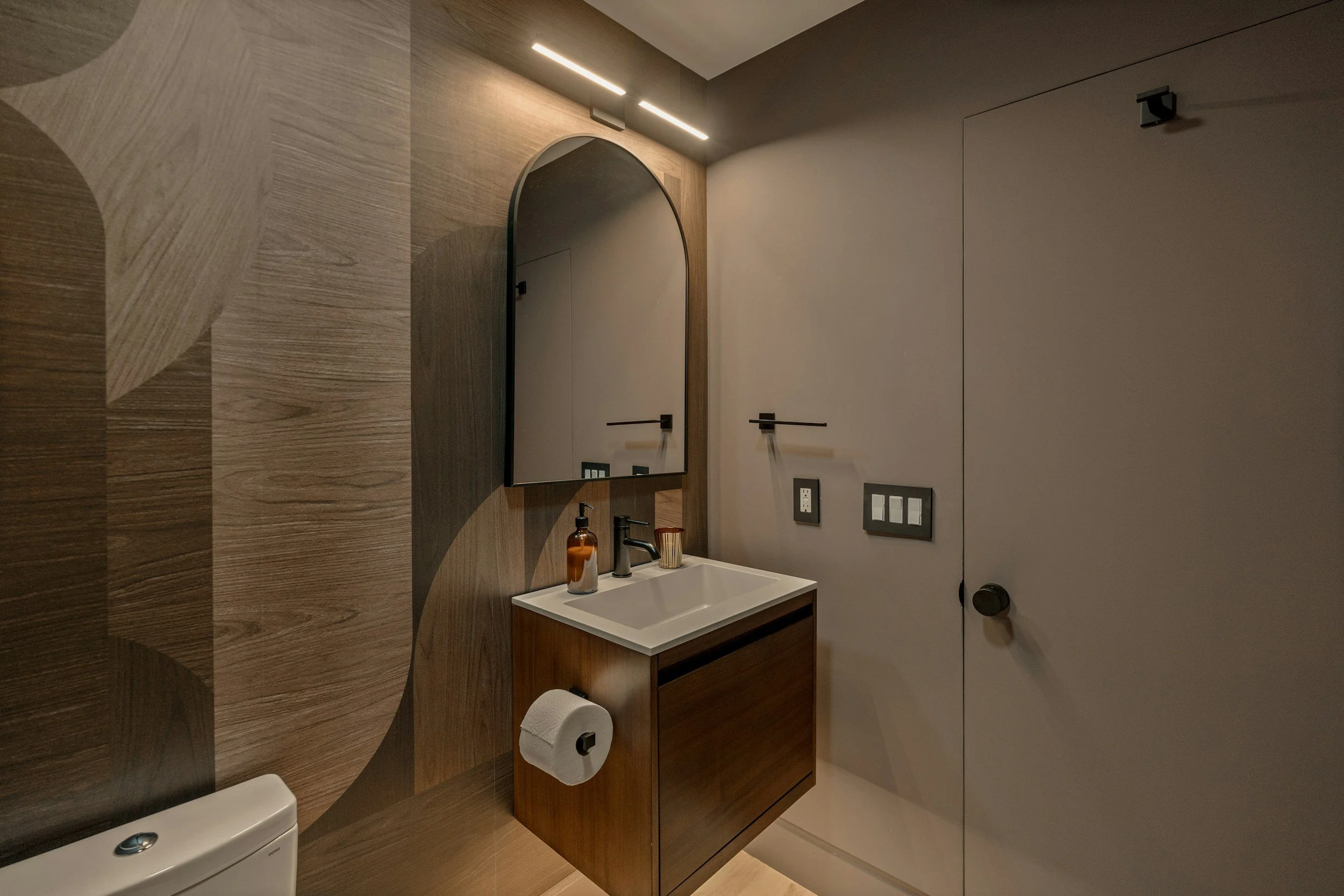 Modern bathroom with wooden wall design, a white sink on a wooden vanity, a mirror, a soap dispenser, a cup, a toilet paper roll, and a door with black handles. There are two light fixtures above the mirror and various electrical panels on the wall.