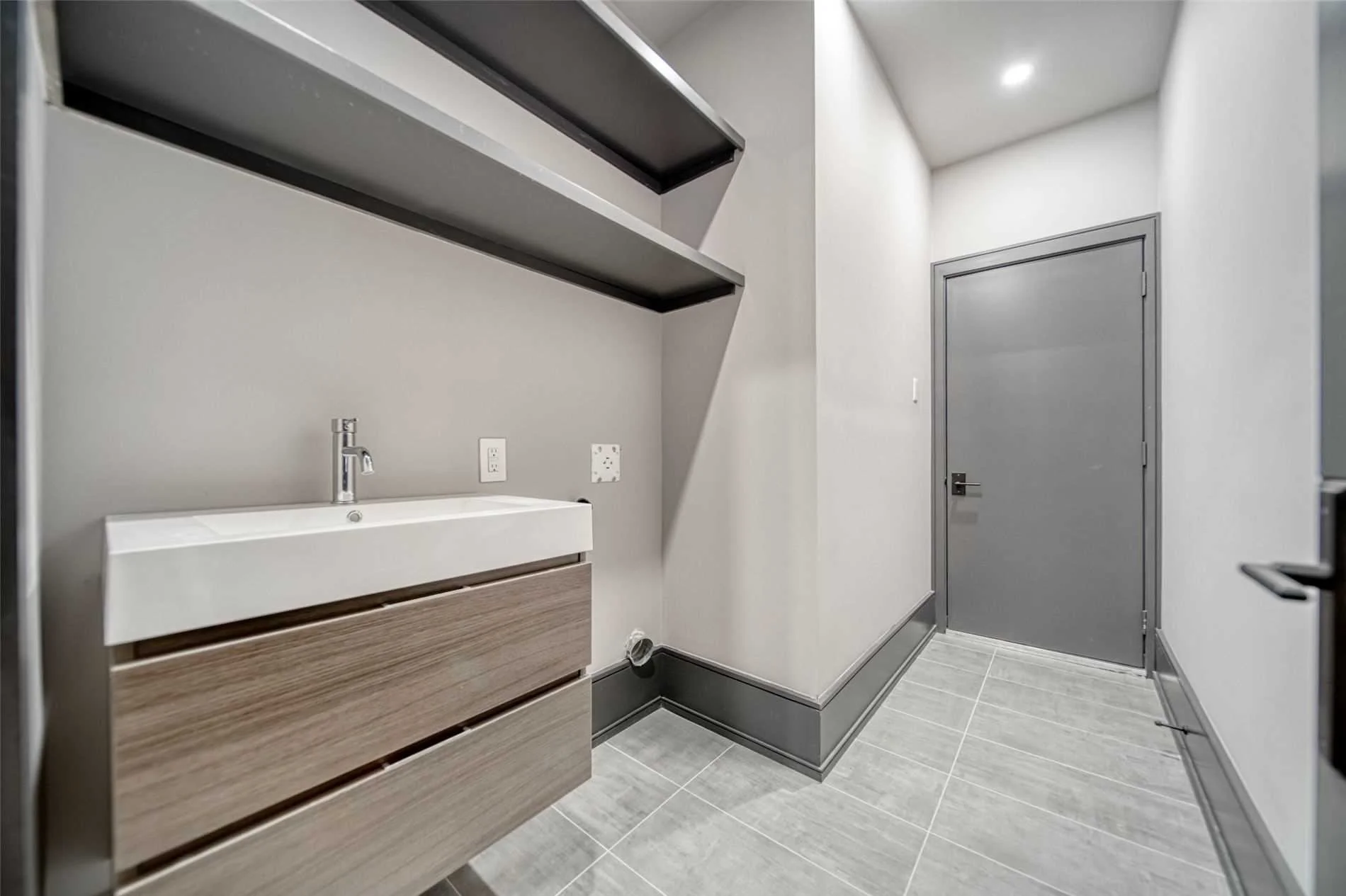 Modern laundry room with gray walls, a white sink and wooden cabinets, gray tiled floor, and gray door.