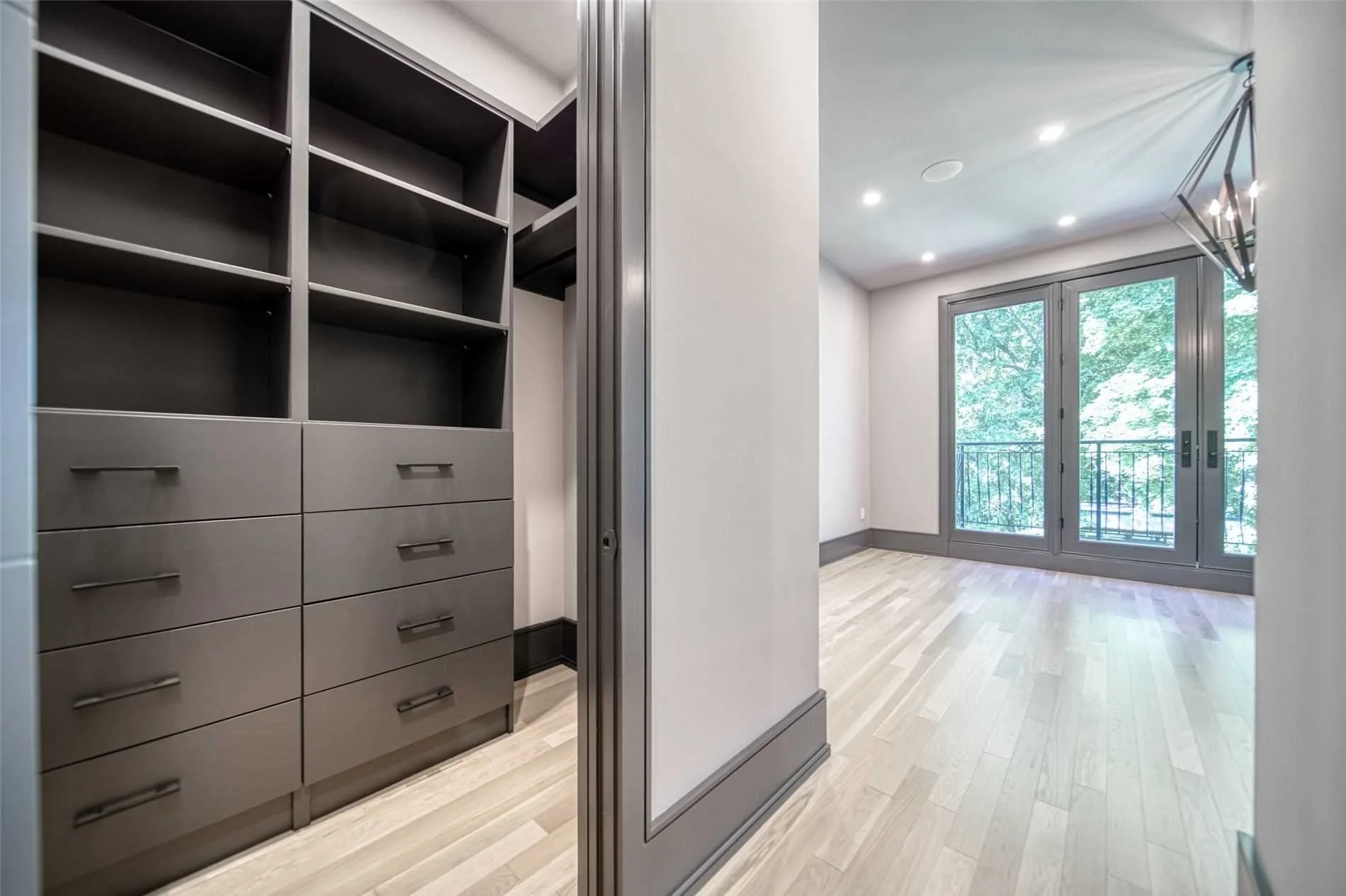 Empty walk-in closet with built-in shelves and drawers, adjacent to a bright room with large windows and a balcony door, hardwood floors, and modern lighting.