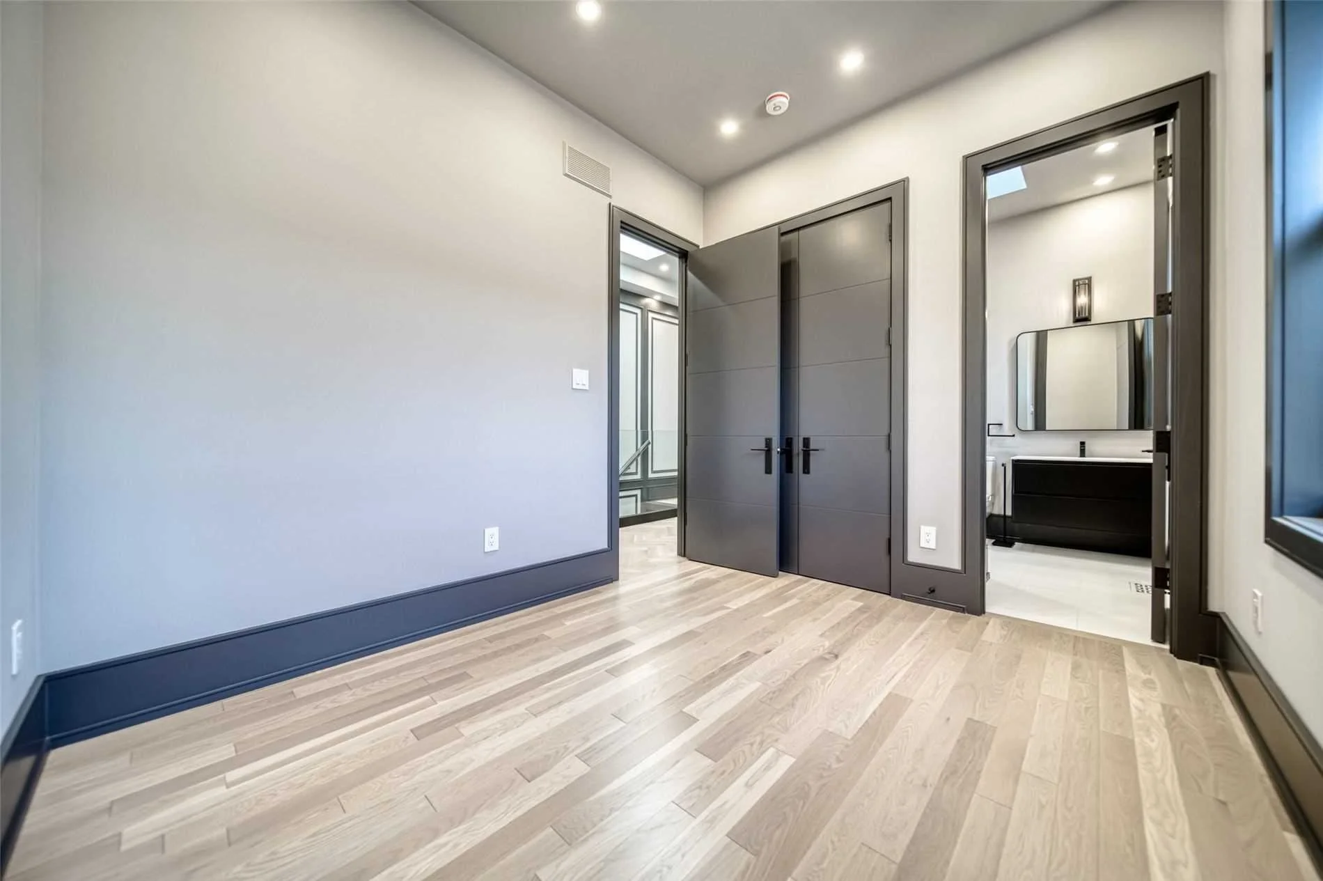 Empty room with hardwood floors, gray doors, and walls, and a view into a bathroom with a large mirror and black vanity.