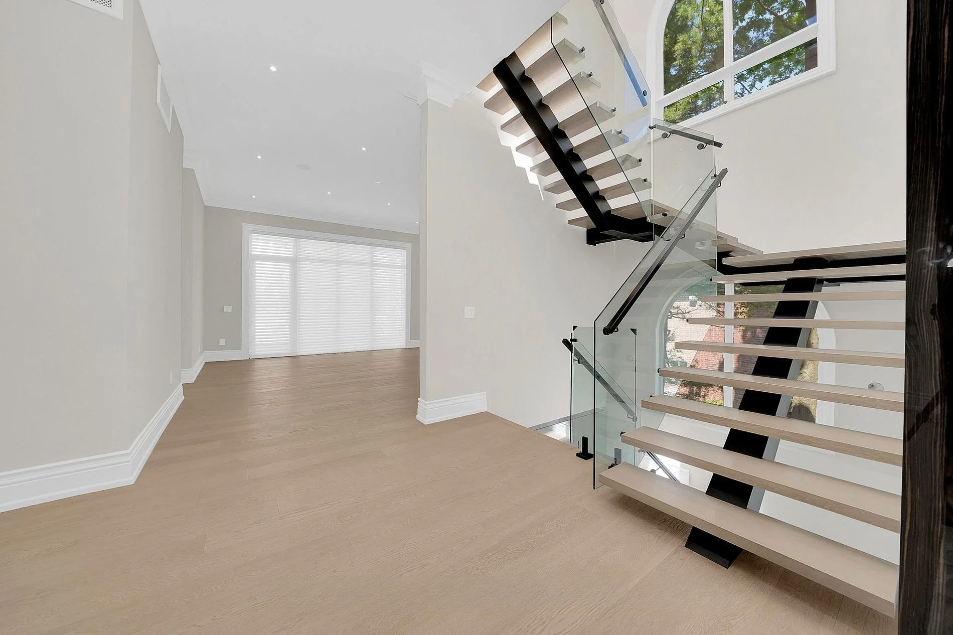 Modern interior with light wooden floors, creamy walls, large windows with white blinds, and a floating staircase with black support beams, glass railing, and open steps, leading to an upper level.