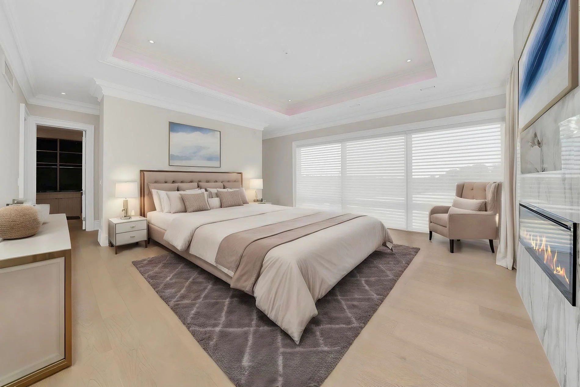 A modern bedroom with a large bed, beige bedding, and multiple pillows. There are nightstands with lamps on either side of the bed, a patterned rug under the bed, and a bay window with blinds. A beige armchair is near the window, and there's a firepl