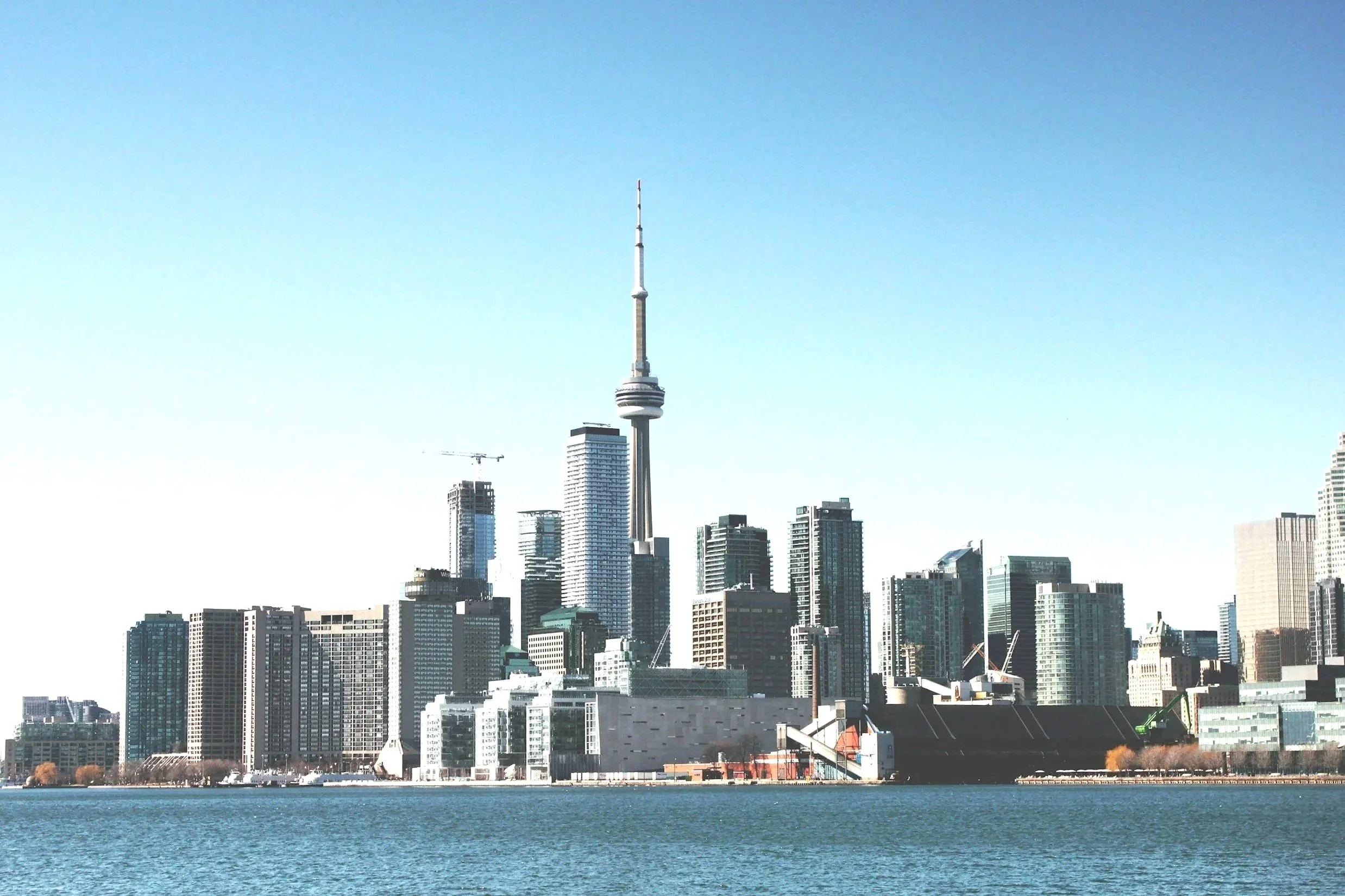 A city skyline of Toronto with the CN Tower prominently in the center, viewed from across a body of water, with clear blue skies overhead.