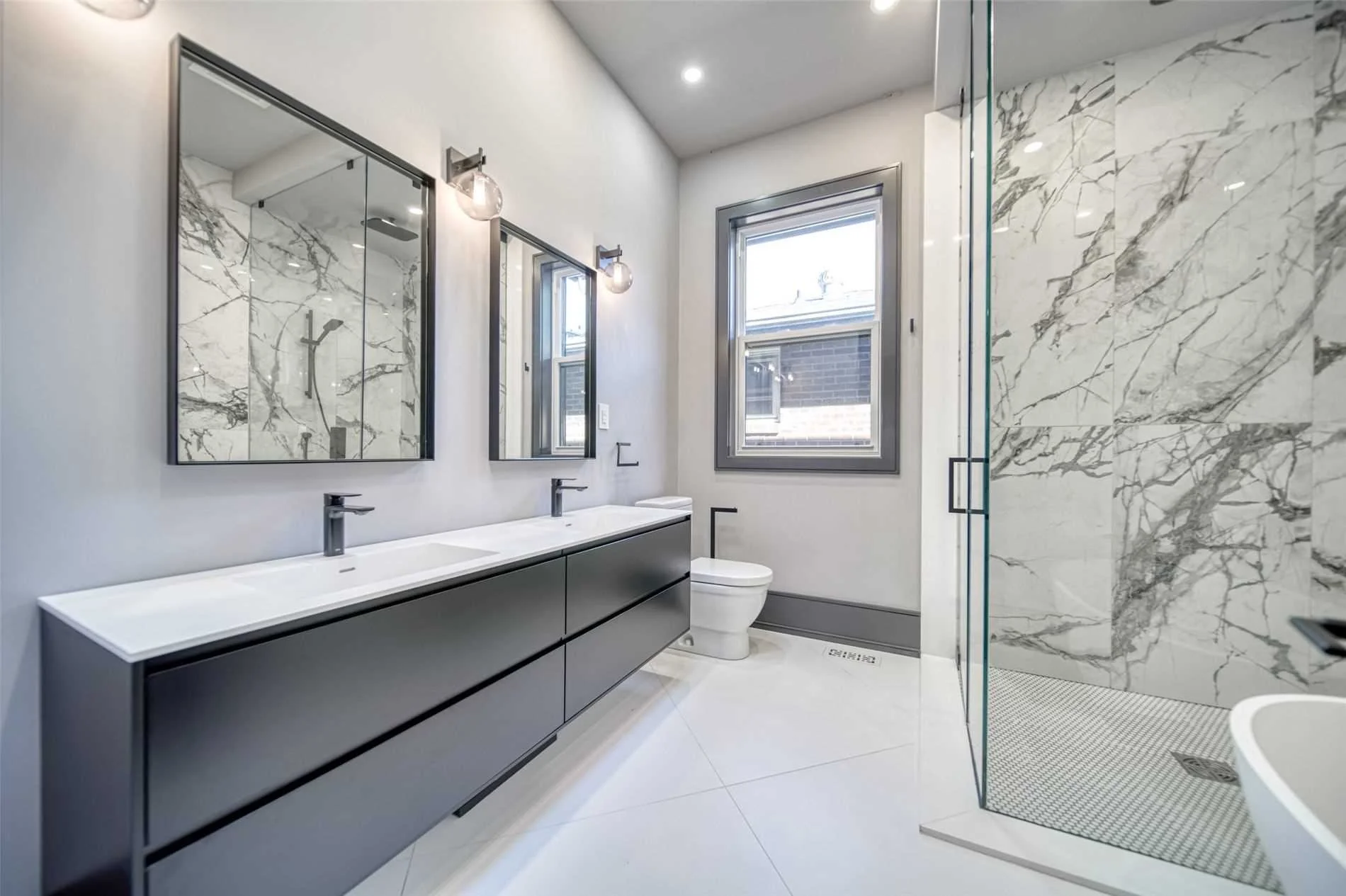 Modern bathroom with double vanity sinks, large mirrors, gray cabinetry, a toilet, a window, and a glass shower with marble walls.
