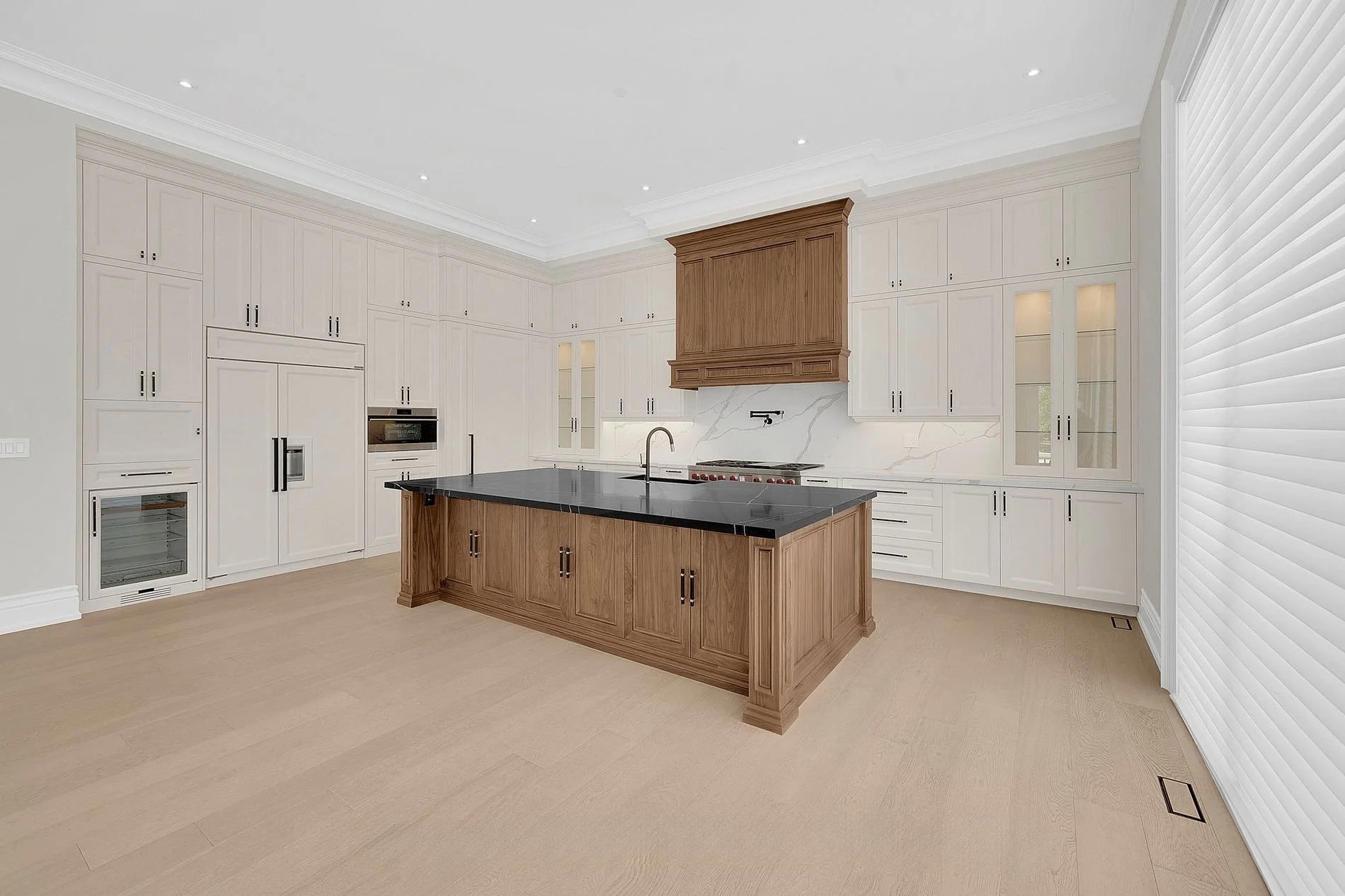 Modern kitchen with white cabinets, a central wooden island with black countertop, and natural light from a large window with white blinds.