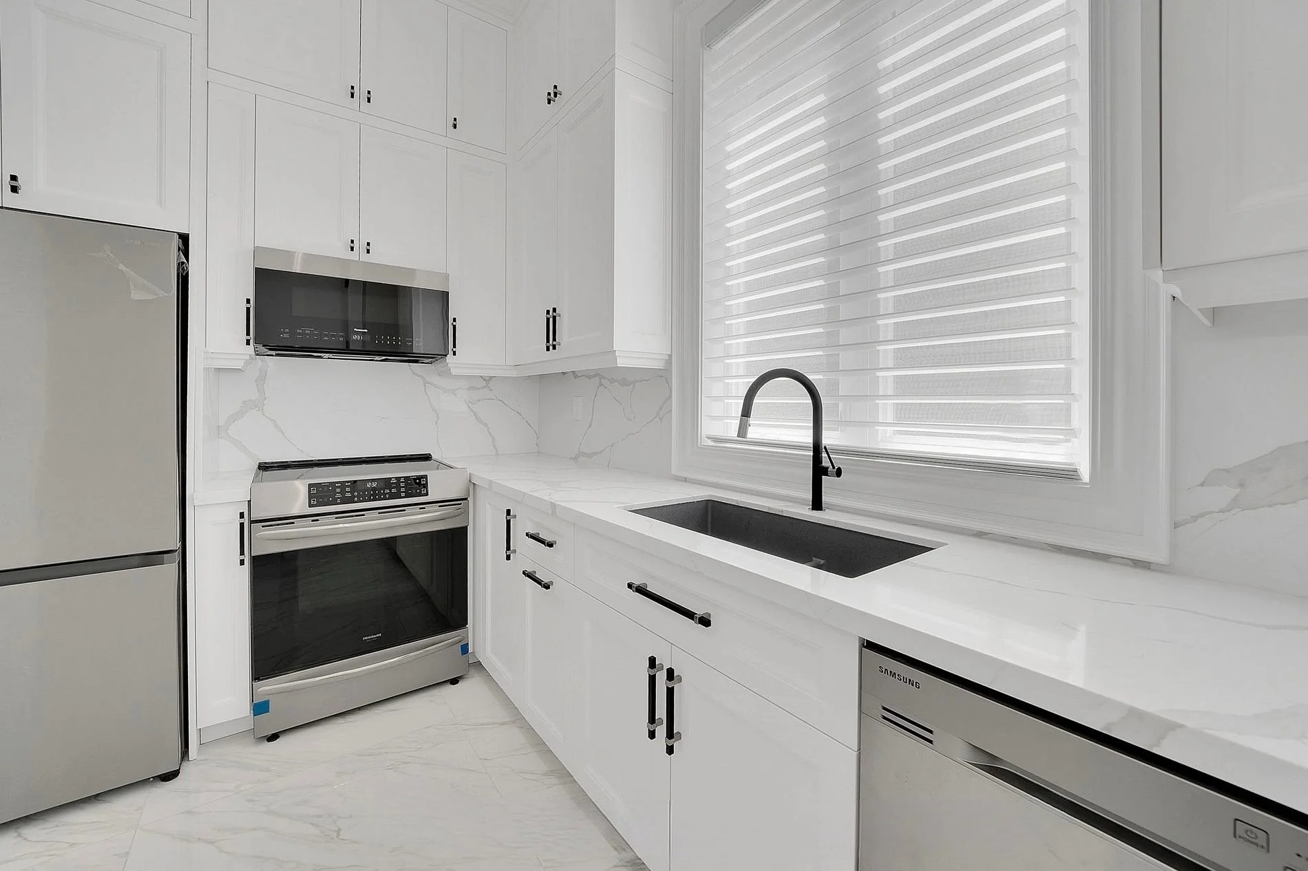 Modern kitchen with white cabinets, marble countertops, black hardware, stainless steel appliances including a refrigerator, oven, microwave, and dishwasher, a black sink, and a large window with white blinds.
