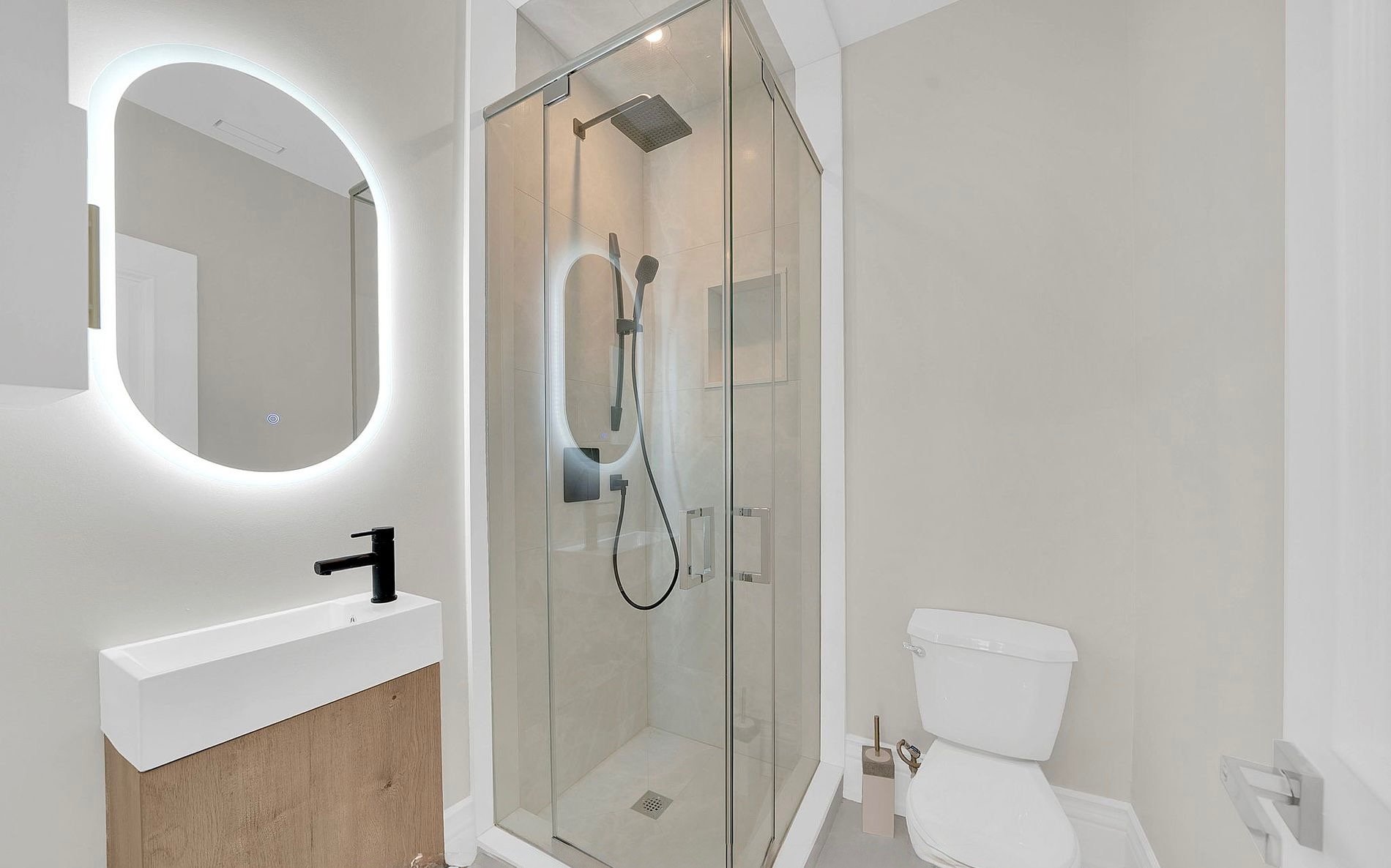 Modern bathroom featuring a glass shower enclosure with a rainfall showerhead and handheld shower, a white wall-mounted sink with a black faucet, an oval backlit mirror, and a white toilet with a toilet brush and holder nearby.
