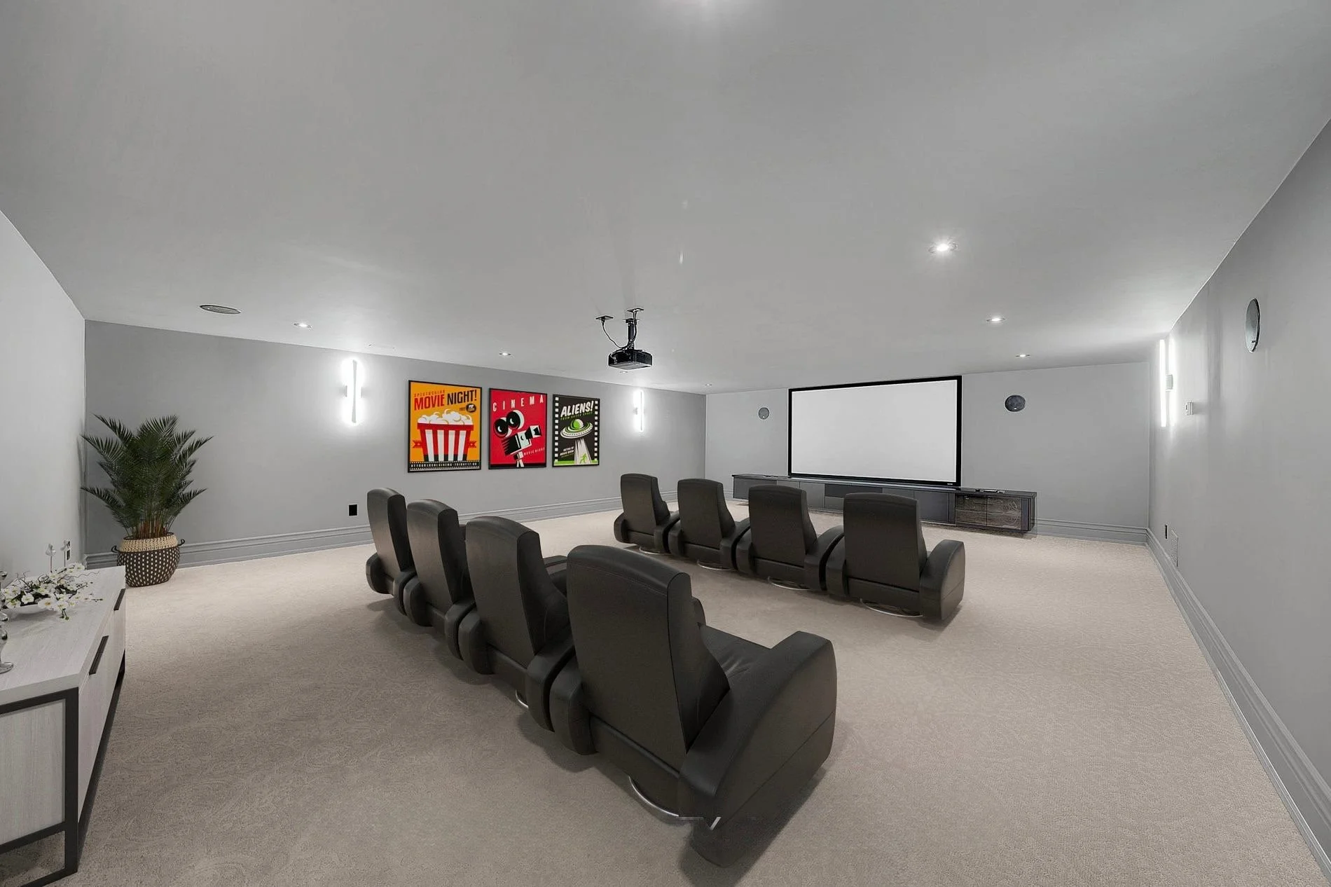 A movie theater room with ten black chairs facing a large blank movie screen. The room has gray walls, a white ceiling, and light-colored carpet. There are three colorful movie-themed posters on the wall, a potted plant, a small table with flowers, a