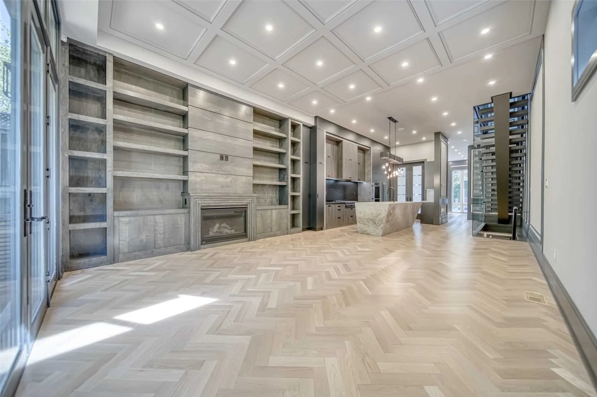 Open-concept living space with light wood herringbone flooring, built-in shelves, a fireplace, and modern kitchen with island and pendant lighting.