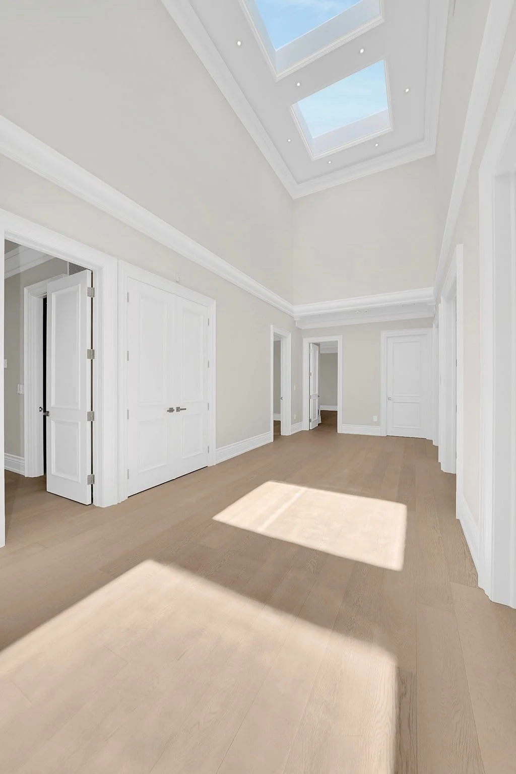 Empty room with white walls, beige flooring, multiple white doors, and skylights on the ceiling allowing natural light in.