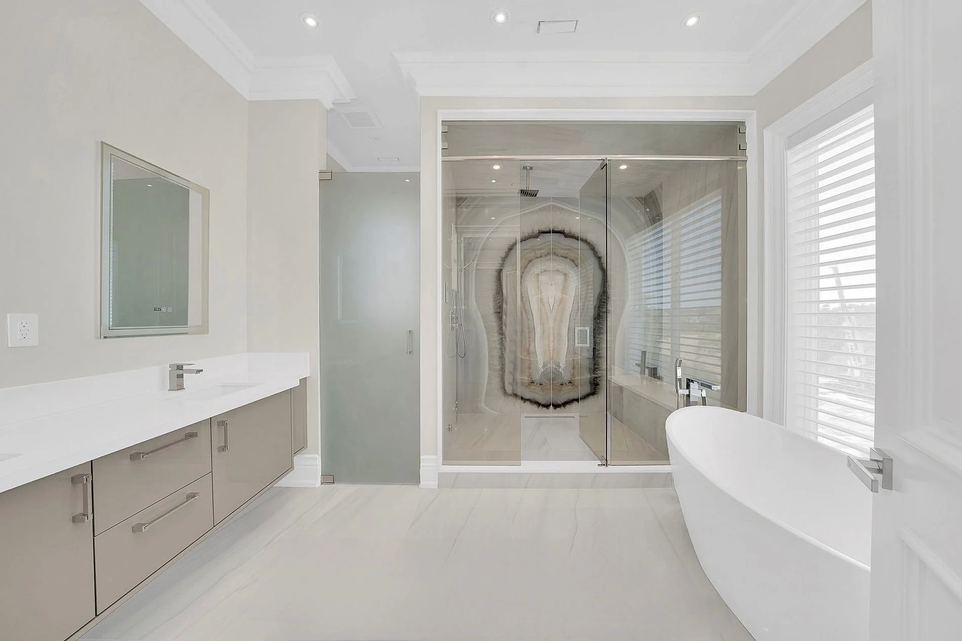 Modern bathroom featuring a walk-in shower with a geode-patterned glass door, a white freestanding bathtub, large windows with white blinds, and a vanity with a mirror and gray cabinet.