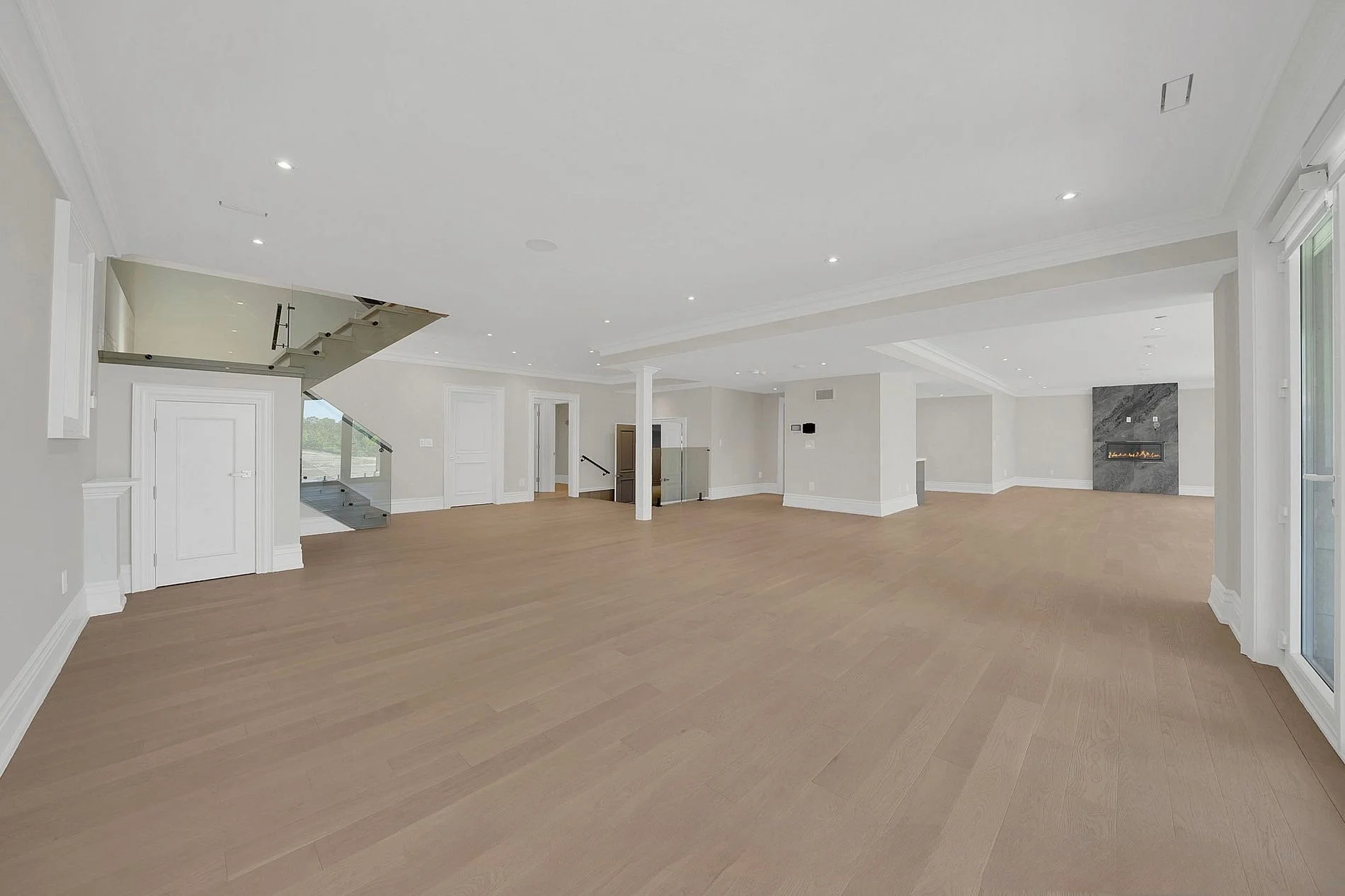 Empty living room with light wood flooring, white walls, modern built-in fireplace, large windows, and a staircase with glass railing.