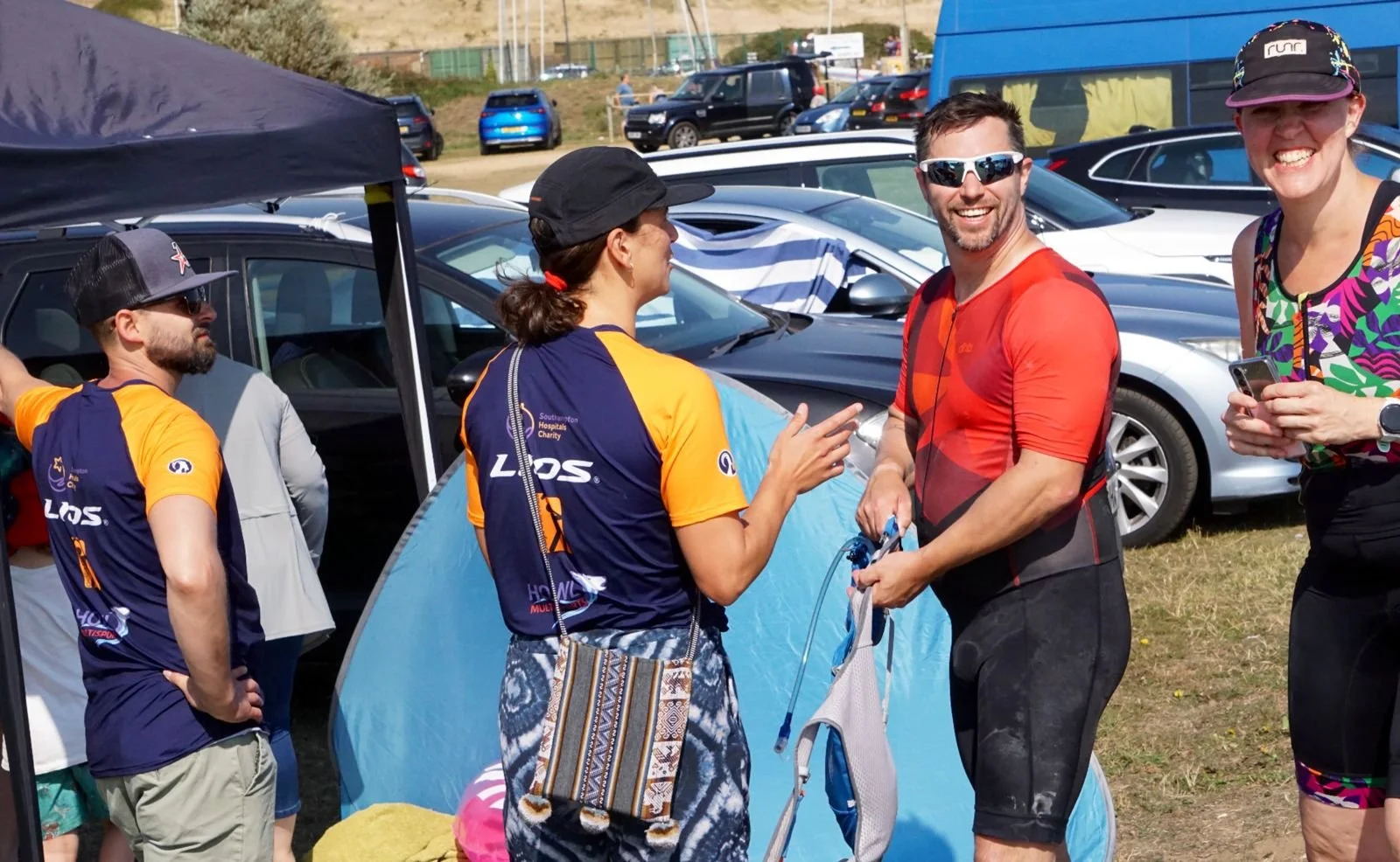 People gathered outdoors near parked cars, some in athletic gear, smiling and talking, with a small blue tent in the background on a sunny day.