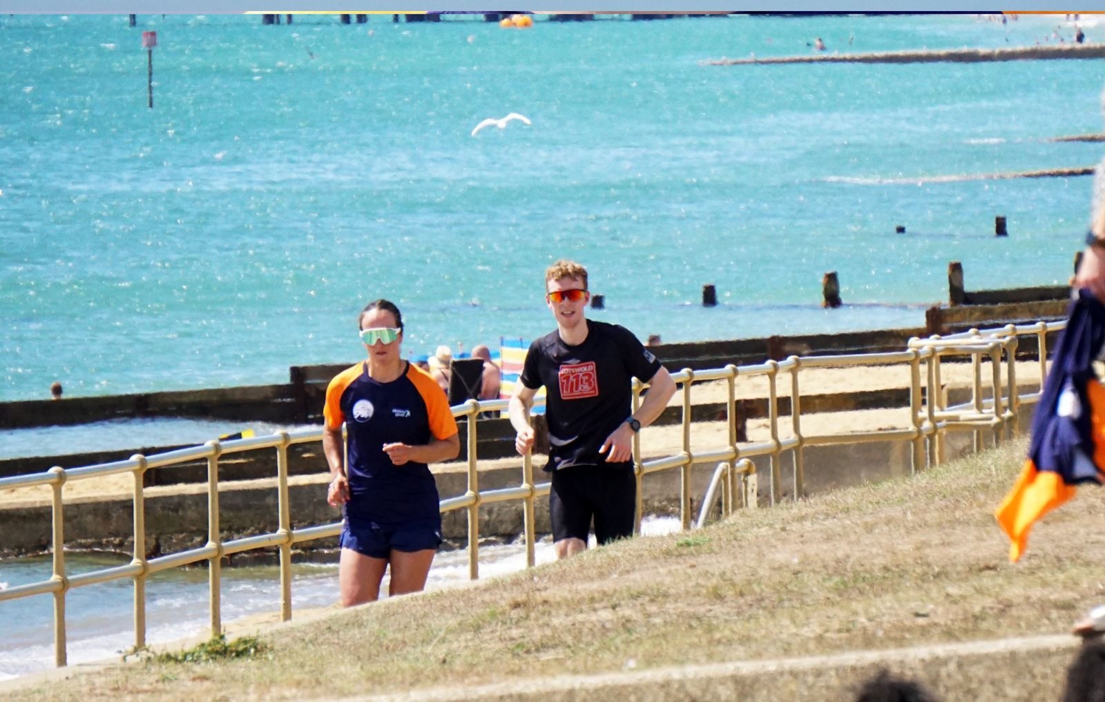 Two runners, a woman and a man, jogging along a beachside path with water and a seagull in the background. The woman is wearing sunglasses, a dark blue and orange athletic shirt, and shorts. The man has red hair, sunglasses, a black shirt with a race
