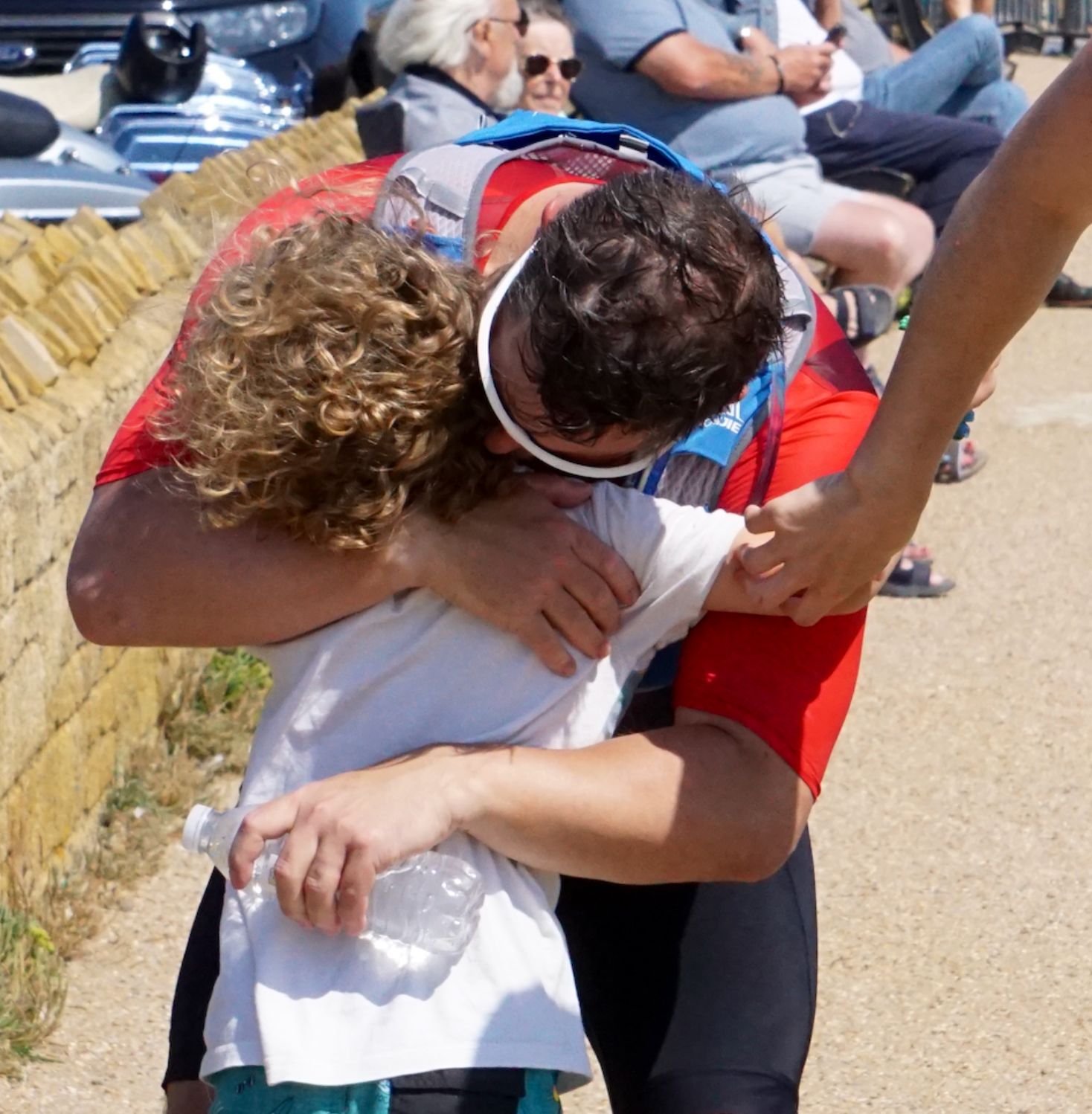 An emotional moment of a person in a red athletic shirt helping a child in a white shirt after what appears to be a sporting event, with other people sitting on a bench in the background.