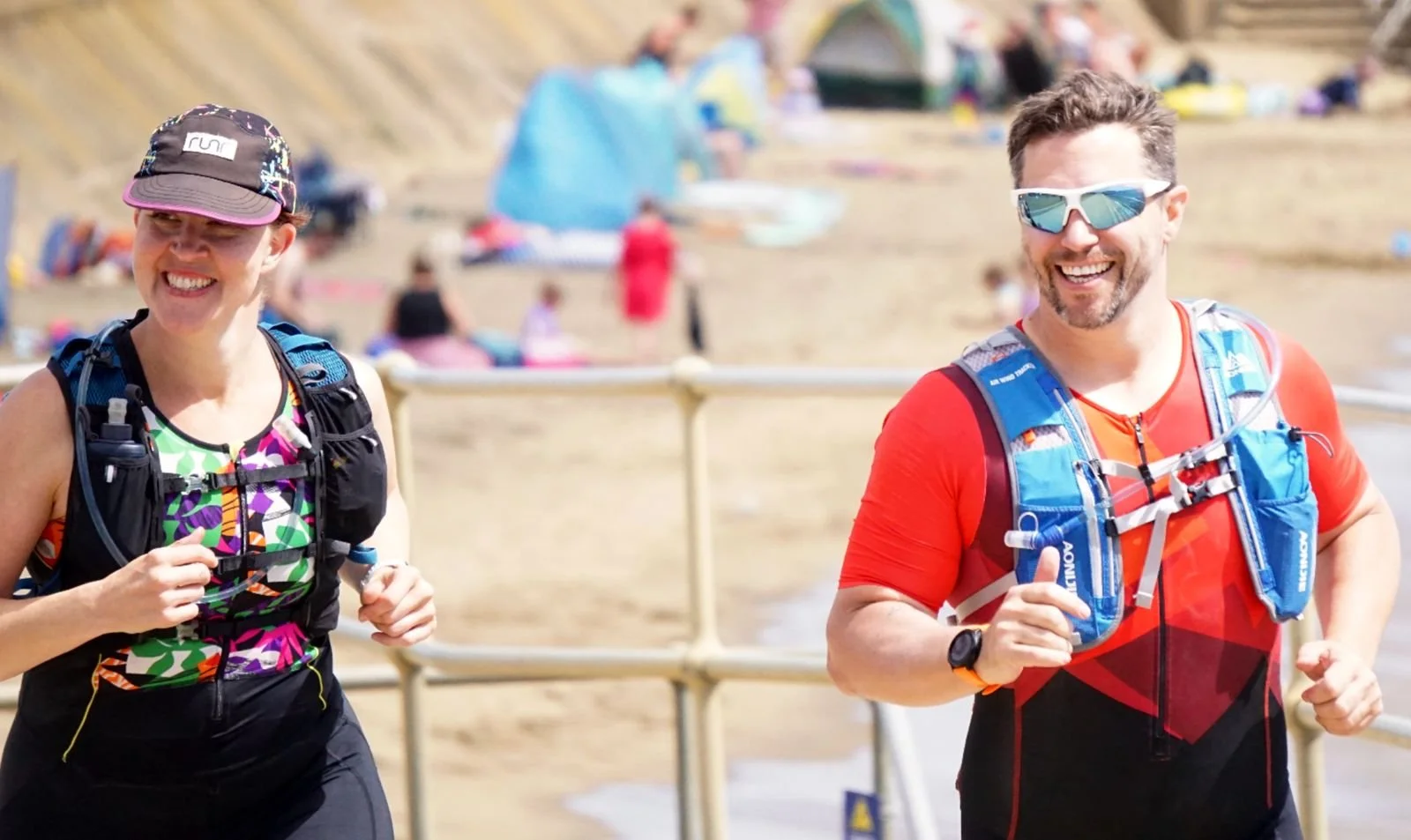 Two people running outdoors on a sunny day, equipped with running gear and hydration packs, smiling and enjoying the activity.