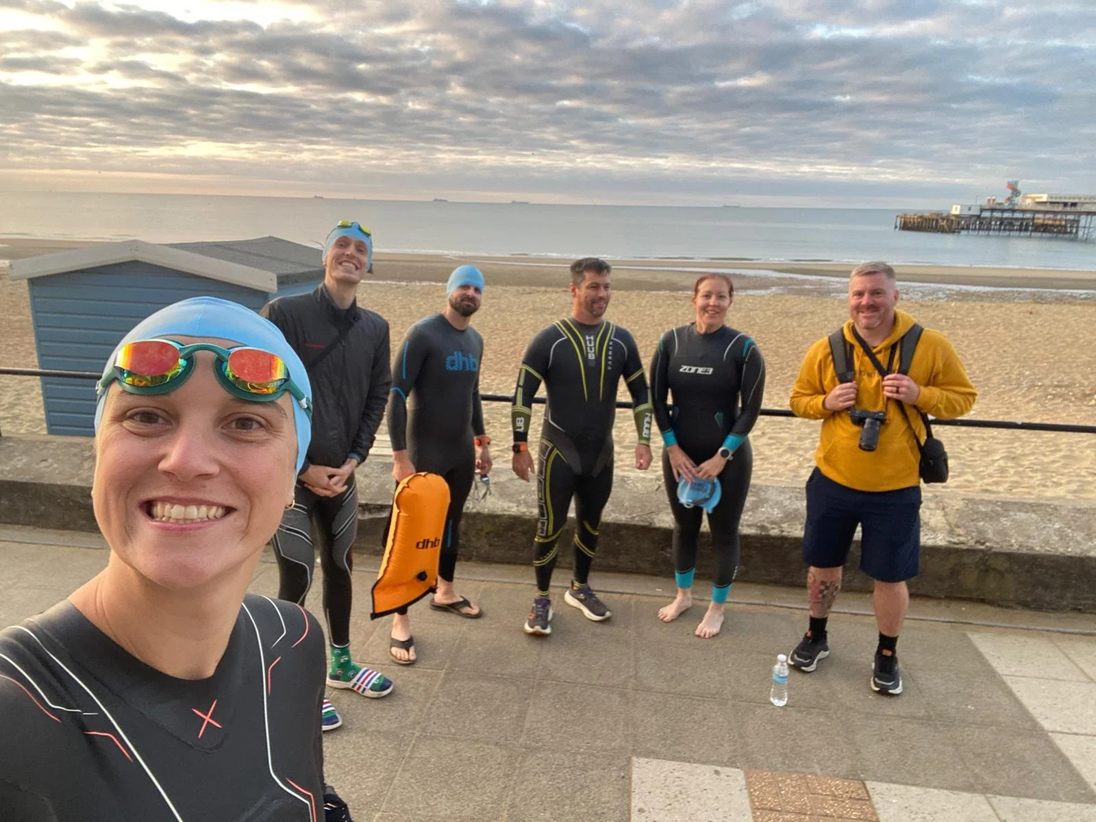 Smiling woman taking selfie with five people in wetsuits at the beach during sunrise or sunset, with ocean, pier, and cloudy sky in the background.