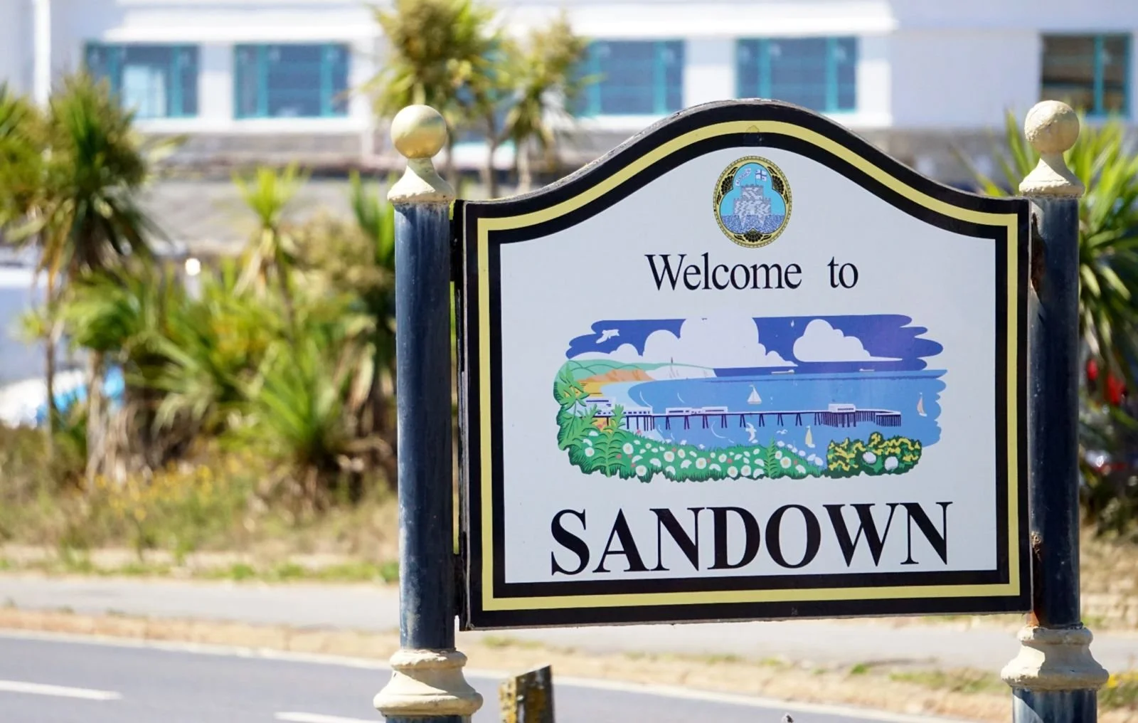 Welcome to Sandown sign with a colorful illustration of a seaside scene, including a pier, sailboats, waves, and clouds, with greenery and buildings in the background.