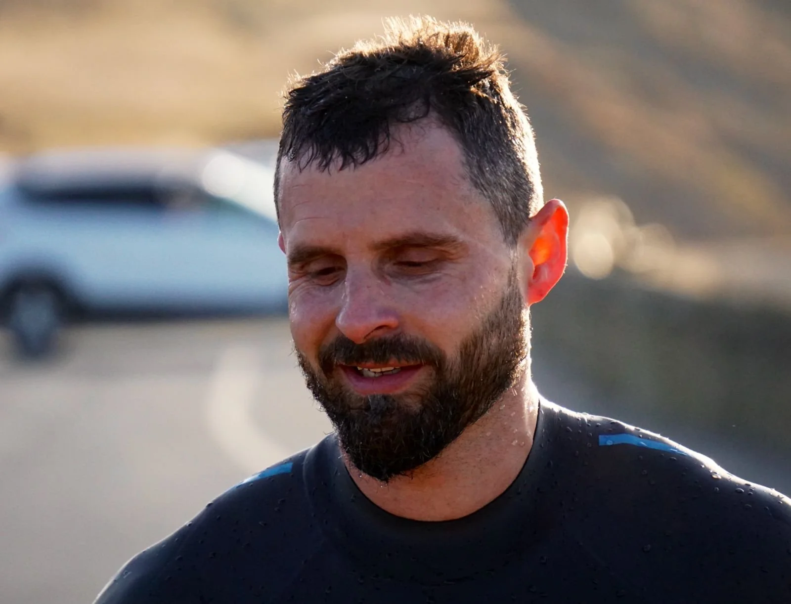 A man with wet hair and a beard, wearing a wet black shirt, smiling slightly, outdoors with a blurred background of a road and a car.