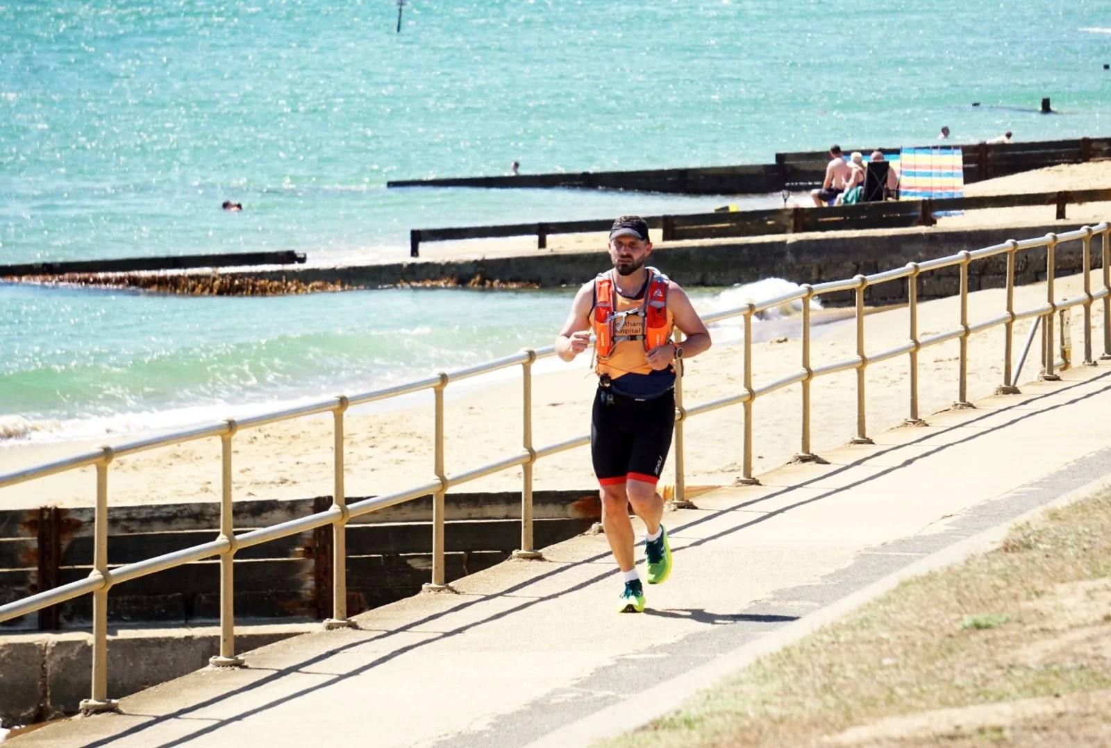 A man jogging along a paved walkway by the beach, with the ocean and beachgoers in the background.