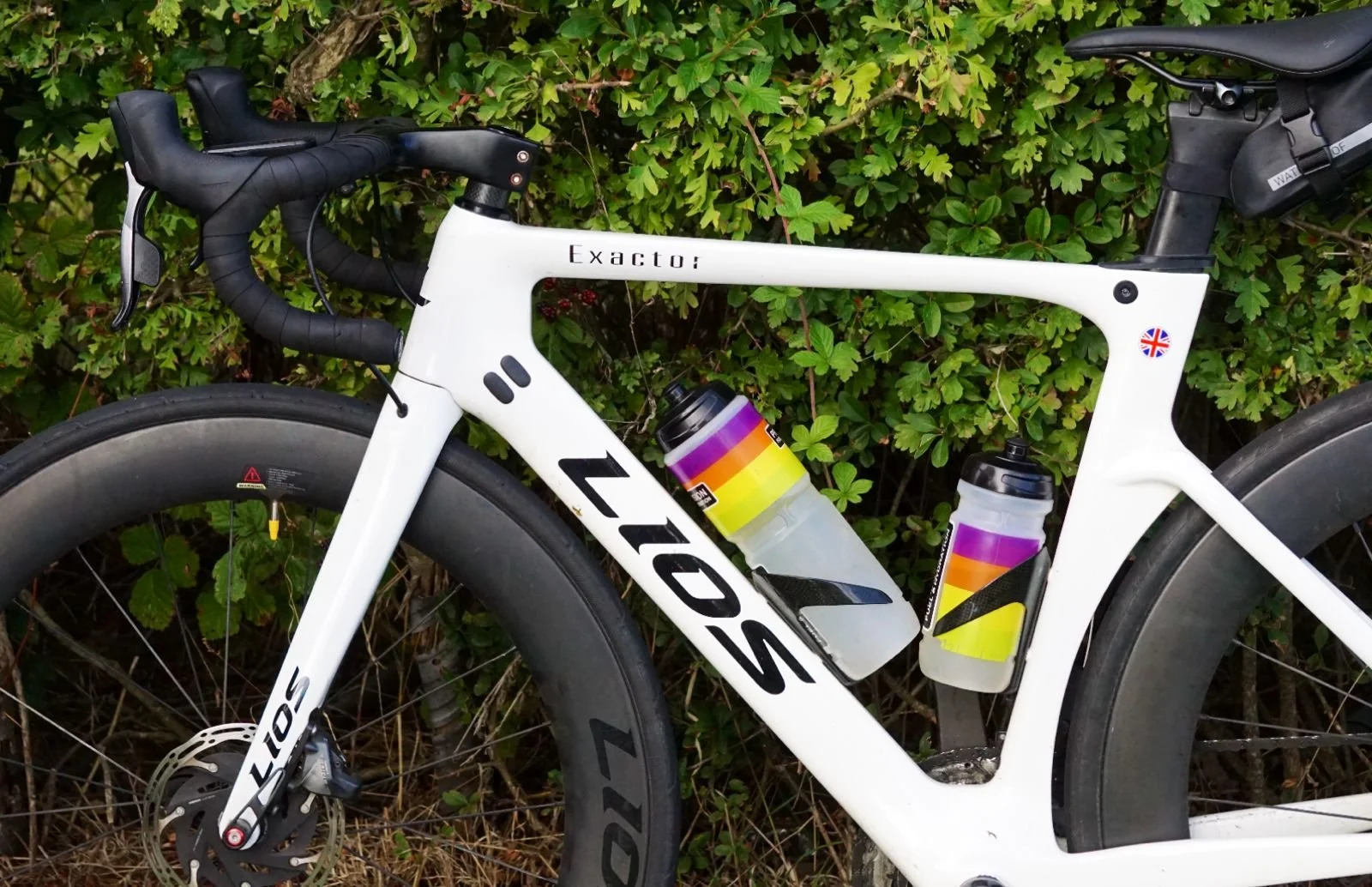 Close-up of a white LIOS road bicycle with two water bottles attached, leaning against green bushes.