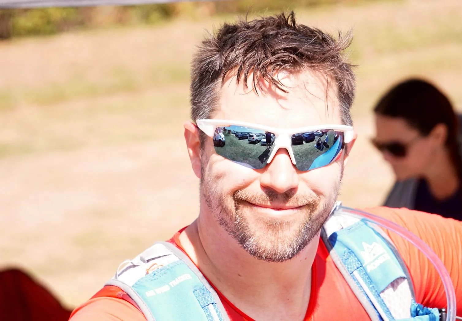 A man smiling at the camera wearing sunglasses with reflecting lenses, an orange shirt, and a light blue and white backpack, with a woman in the background also wearing sunglasses, outdoors on a sunny day.