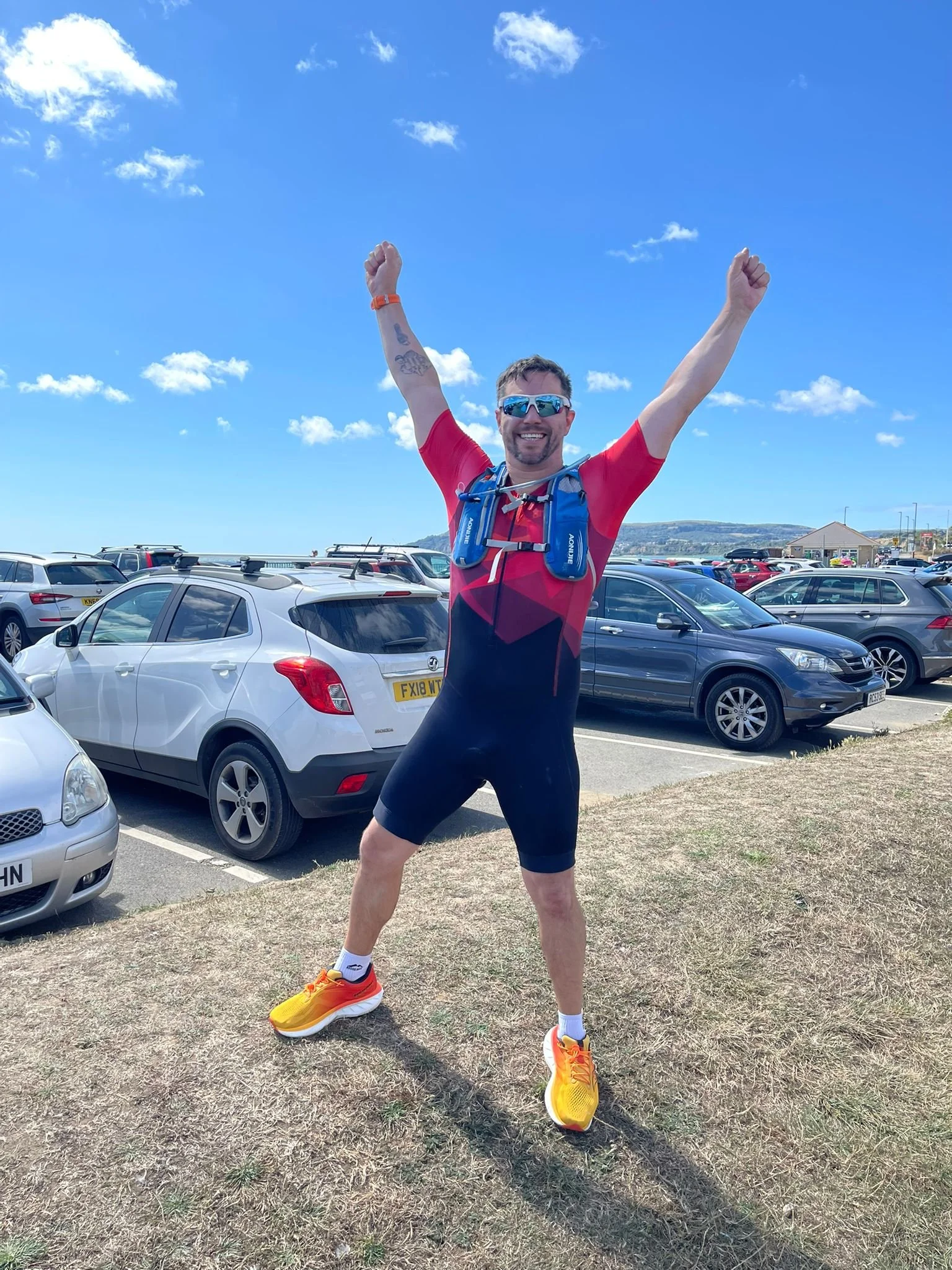 A man in athletic gear outdoors, smiling with arms raised, standing on grass near parked cars, with a blue sky and clouds overhead.