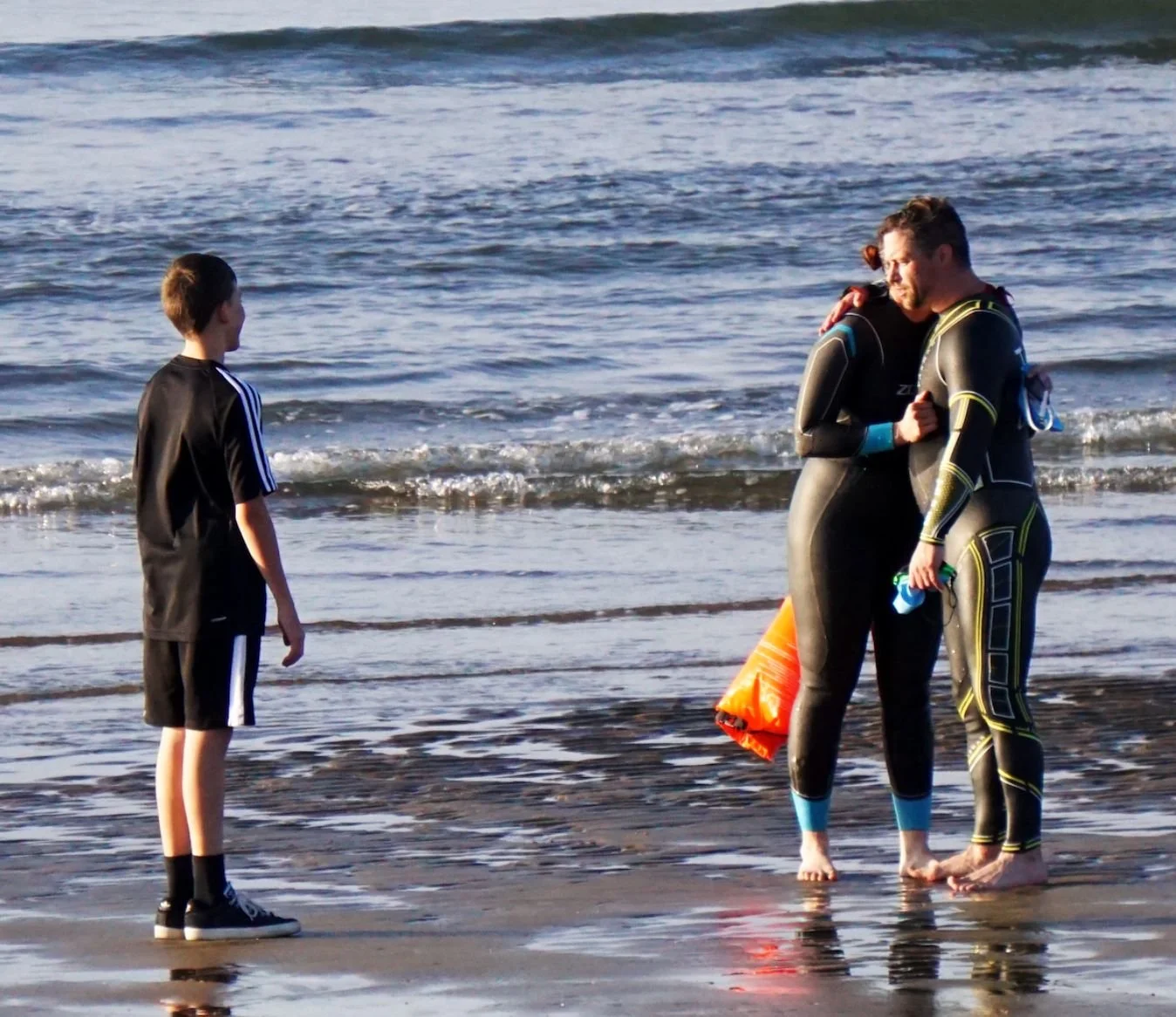 Two men in wetsuits hugging on a beach, a boy stands nearby watching, with ocean waves in the background.