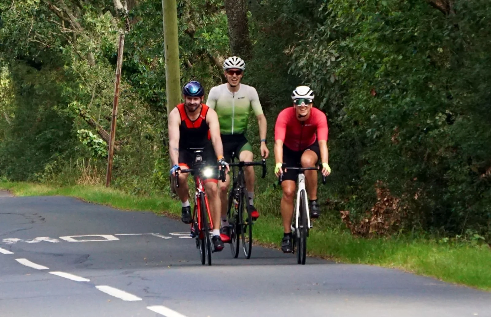 Three male cyclists riding on a road surrounded by trees and green foliage.