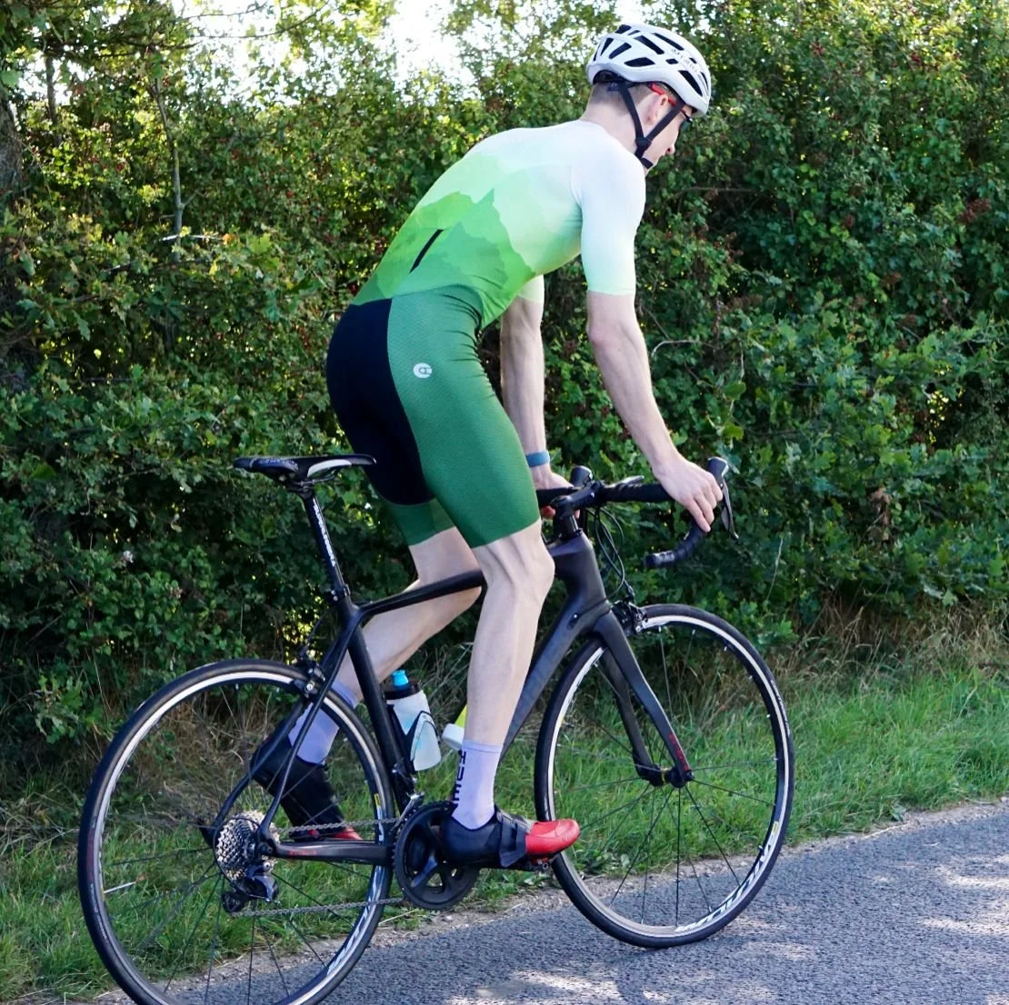 A cyclist riding a black road bike on a paved path with green bushes and trees in the background.
