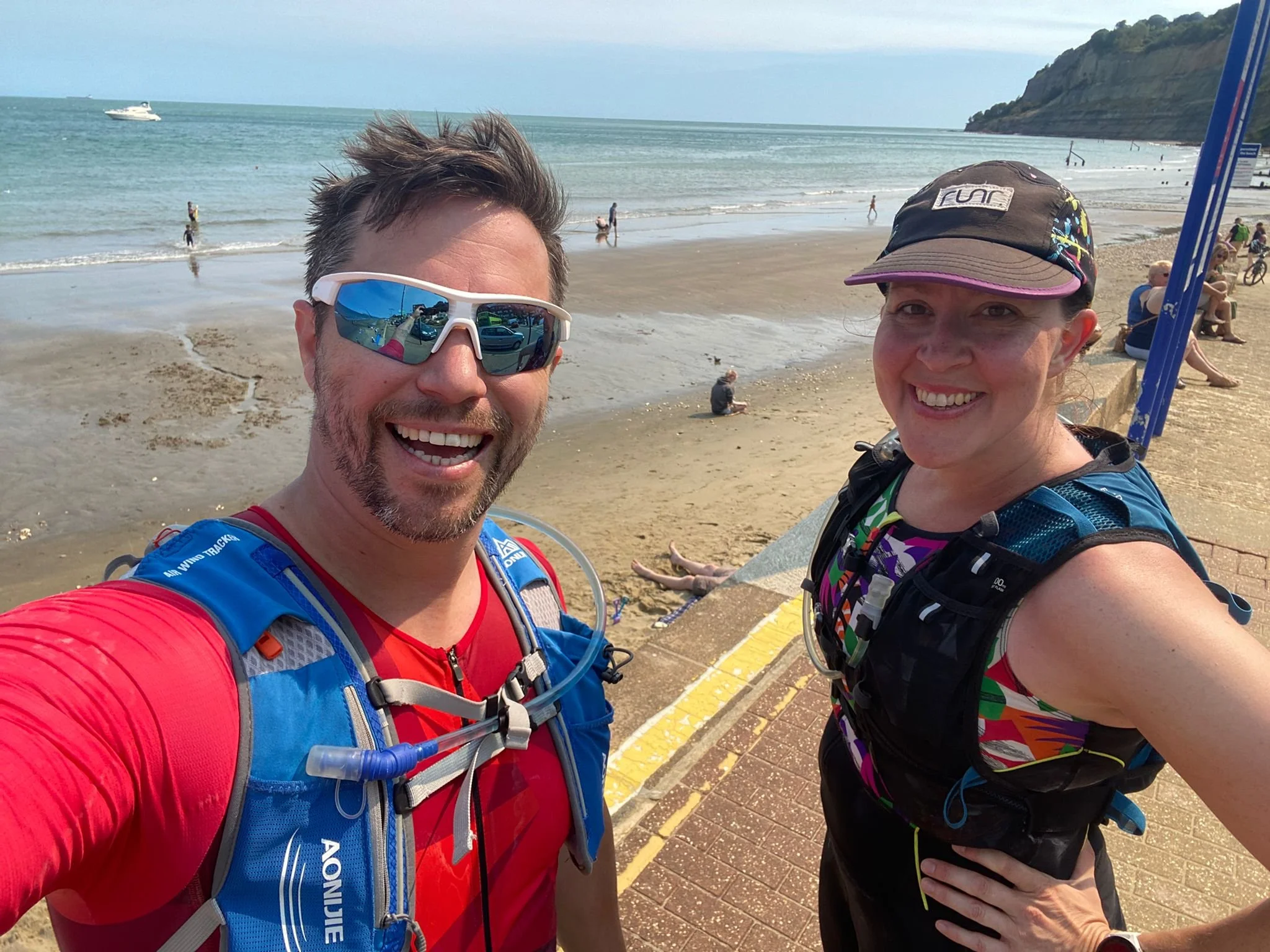 Two smiling runners taking a selfie on a beach, with people swimming and relaxing in the background and a boat in the water.