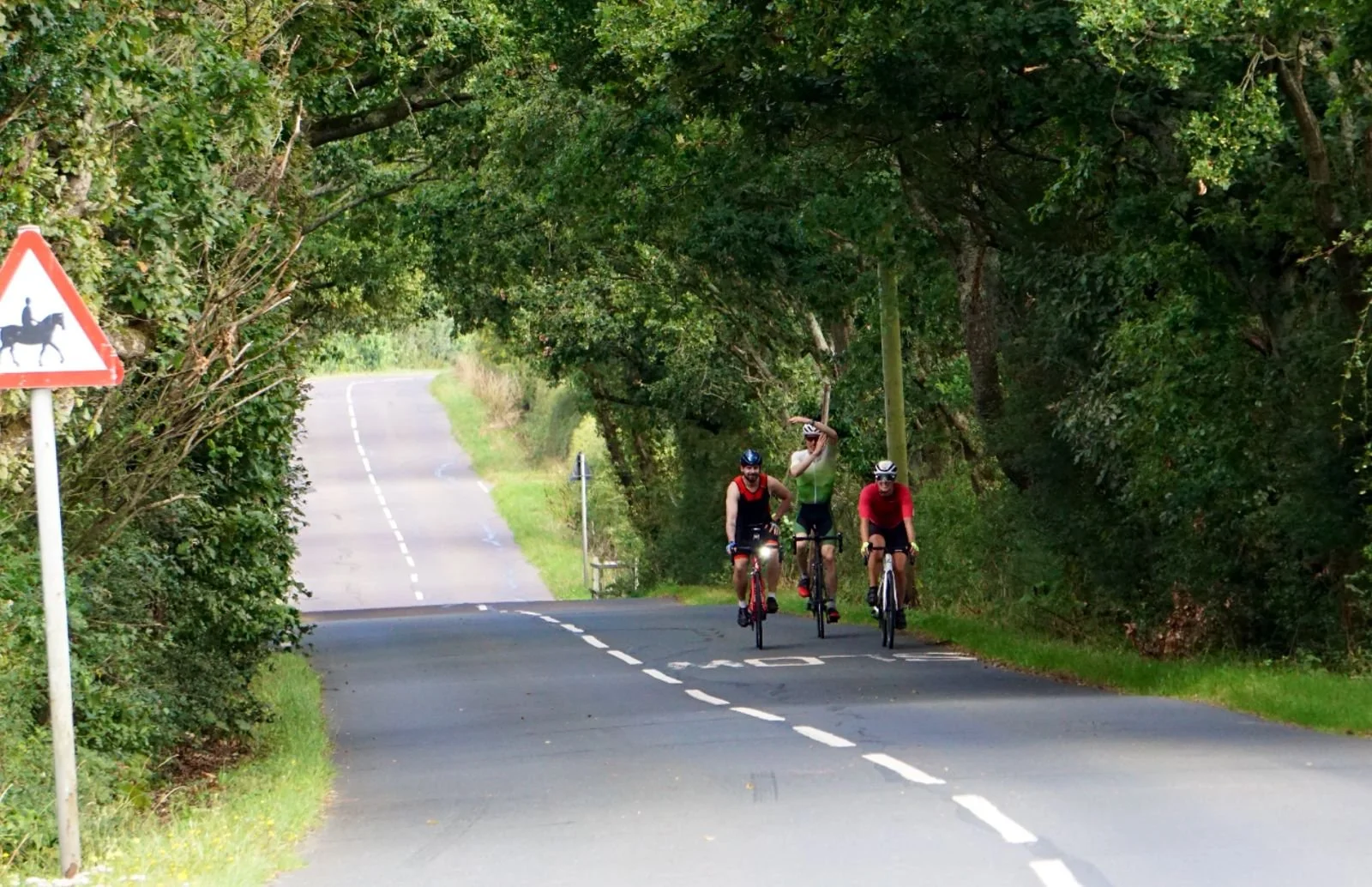 Three cyclists riding on a rural road surrounded by trees, with a warning sign for horseback riders on the left.