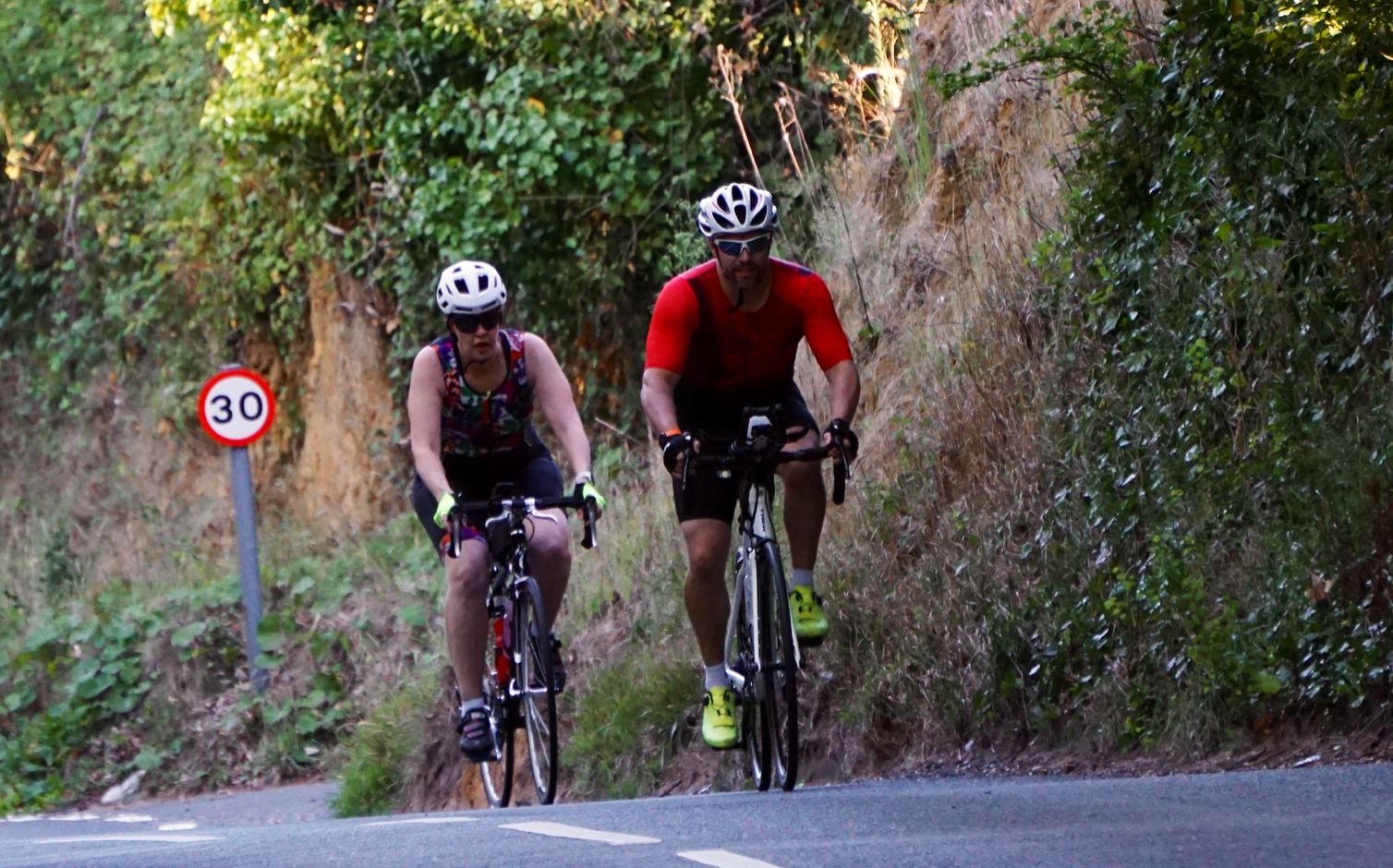 Two cyclists riding uphill on a mountain road through forested area, with a 30 km/h speed limit sign visible on the left.
