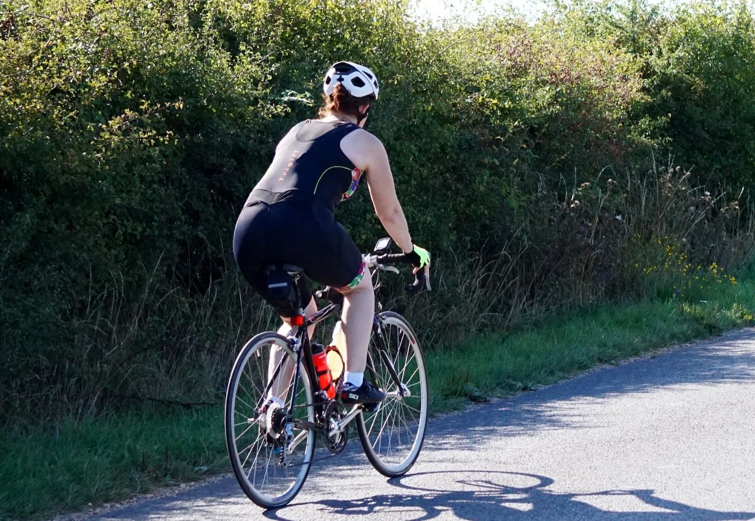 A person riding a road bike along a paved path, wearing a helmet, sleeveless top, and shorts, with a body of bushes and grass on the side.
