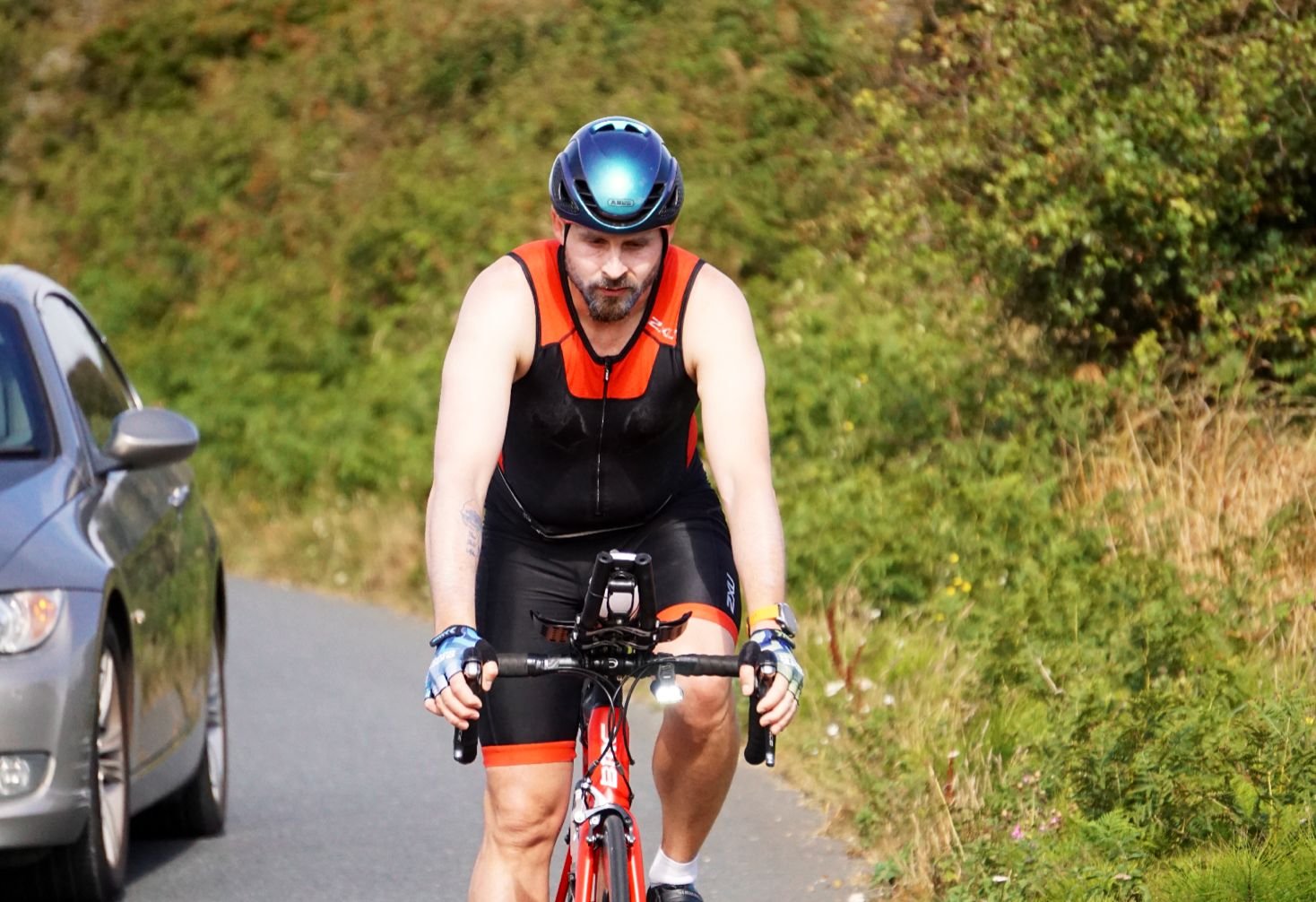 A man riding a bicycle on a rural road with green bushes on the side, wearing a black and orange sports outfit, a blue helmet, and gloves.